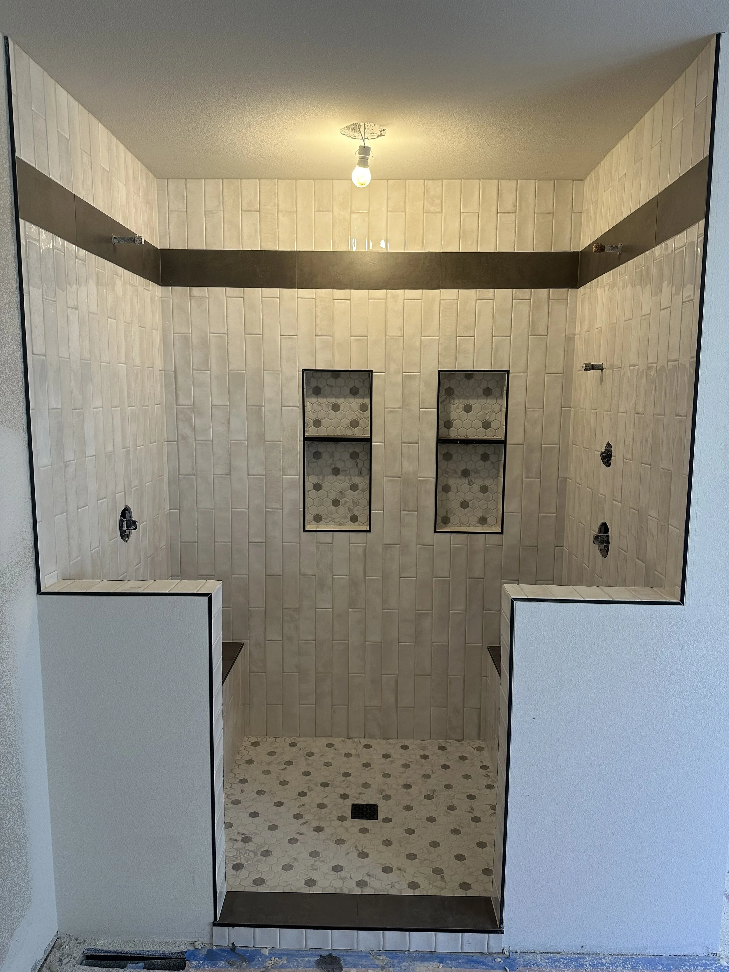 Empty shower with tiled walls and floors, two built-in shelves with hexagonal tile patterns, and a drain in the center of the floor.