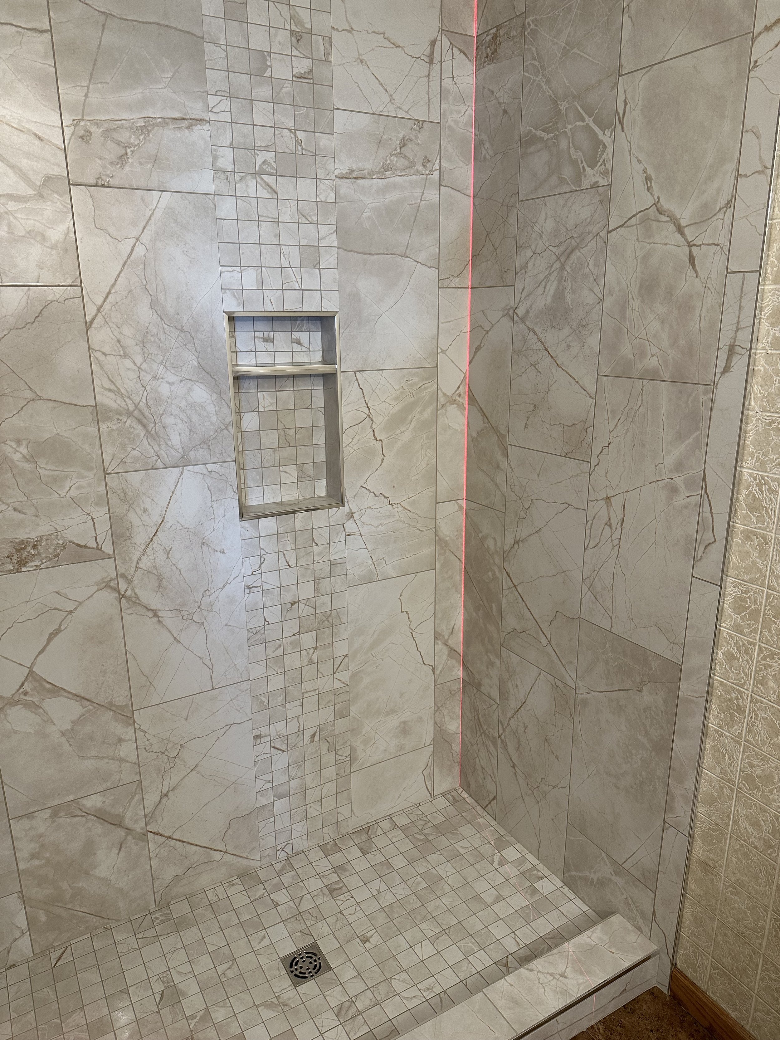 A tiled walk-in shower with beige marble-like walls and a small built-in niche with square tiles. There is a circular drain on the floor, and a red laser level line running vertically along the corner of the shower.