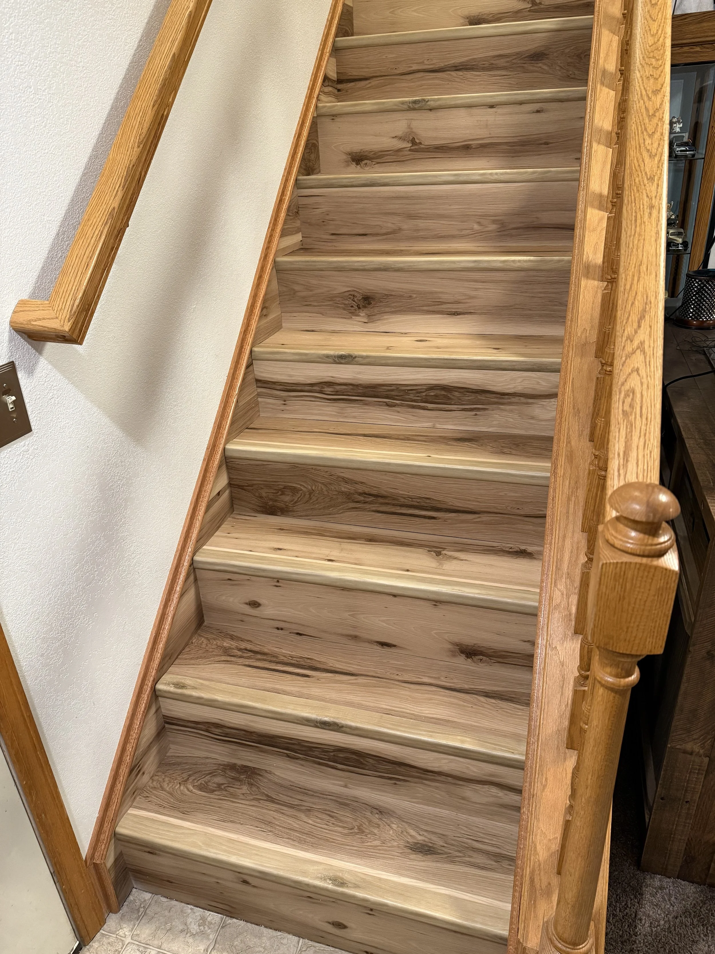 Wooden staircase with natural grain, bordered by a wooden handrail, right next to a white textured wall and a tiled floor.