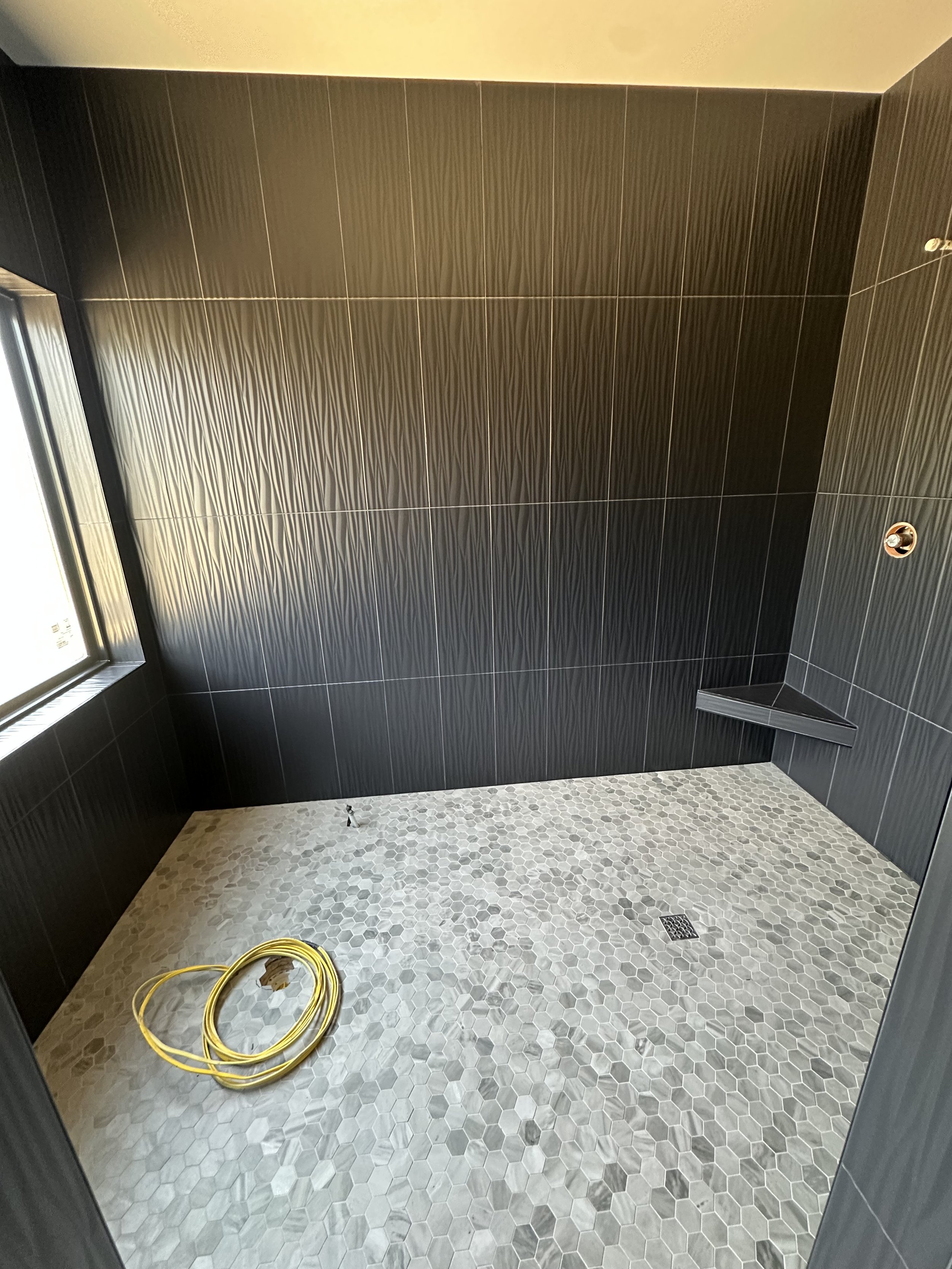 A bathroom with dark tiled walls, a small corner shelf, a drain on the hexagonal tile floor, a window, and a yellow hose on the floor.