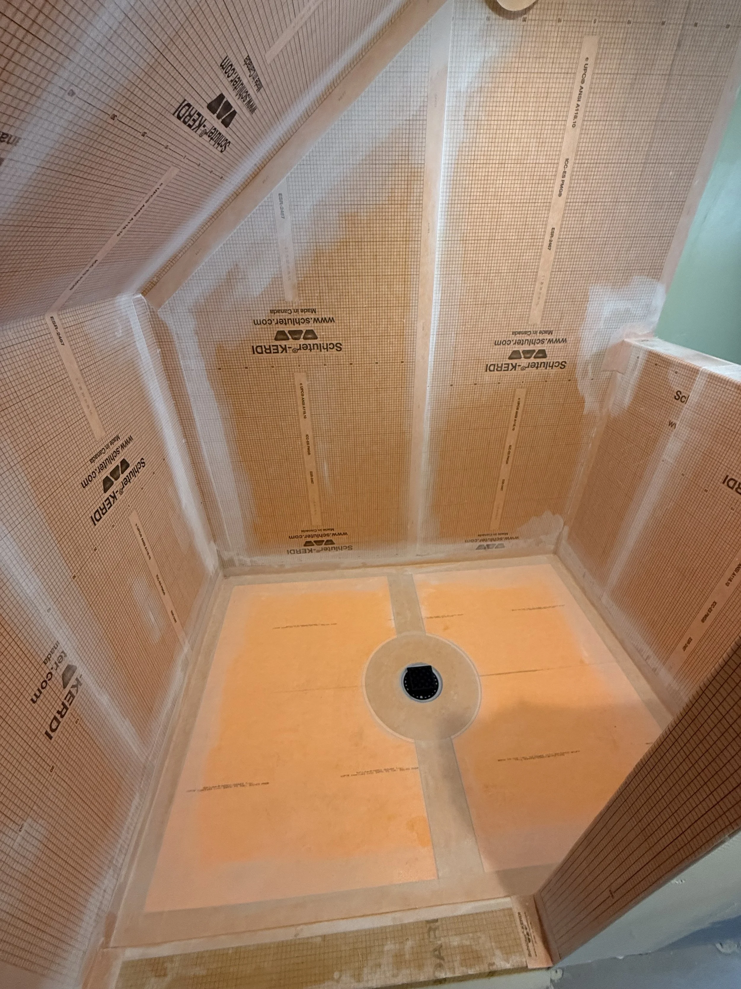 The image shows the interior of a shower with waterproofing membrane installed on the walls and ceiling. The membrane is beige and branded with 'Schluter®-KERDI' and 'Schluter-Systems'. There is a round shower drain at the floor center, and the shower walls have various measurements marked on them for installation.