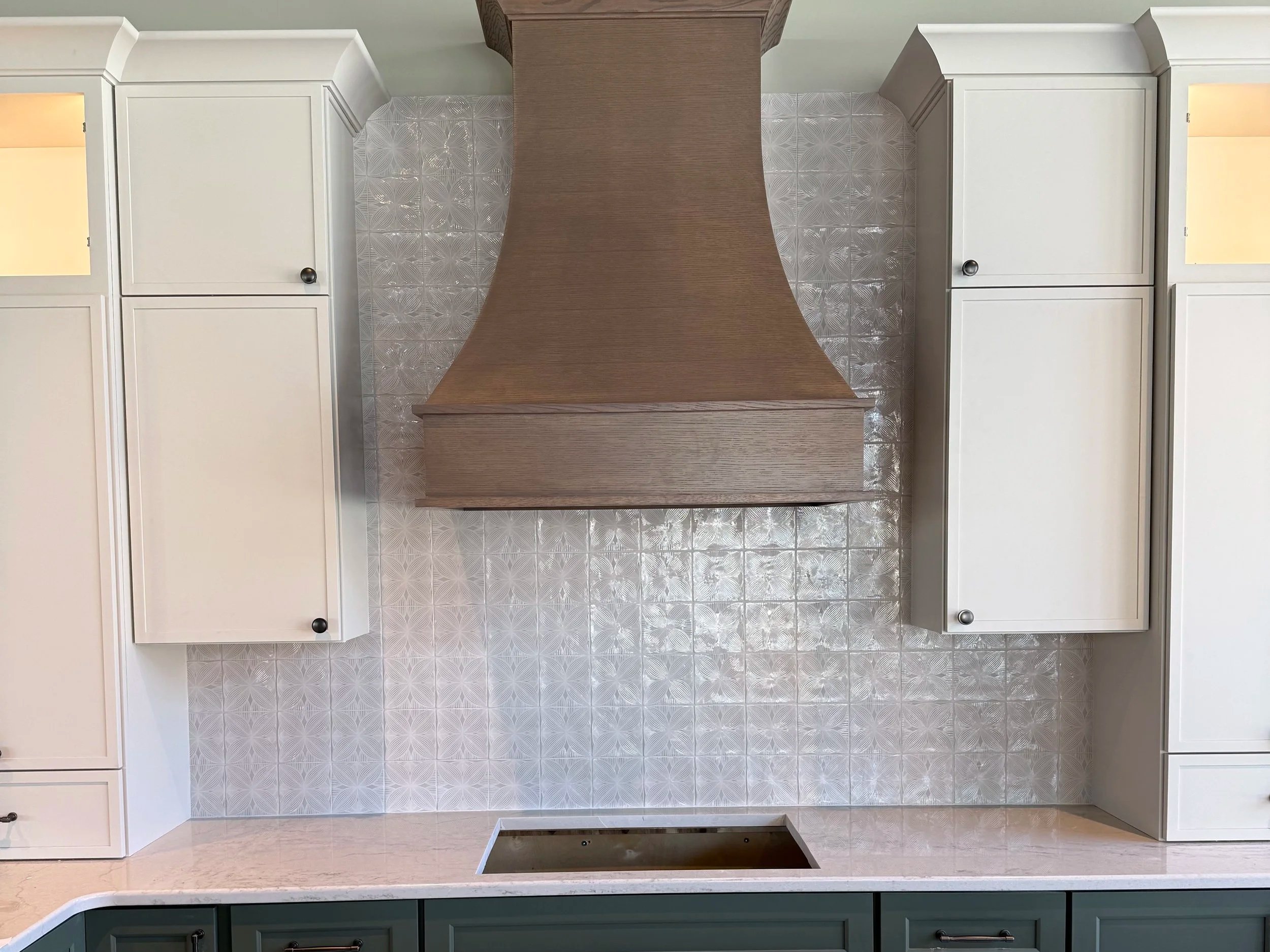 Kitchen backsplash with intricate geometric pattern, wooden range hood, white cabinets with metallic knobs, and a white countertop with a built-in sink.