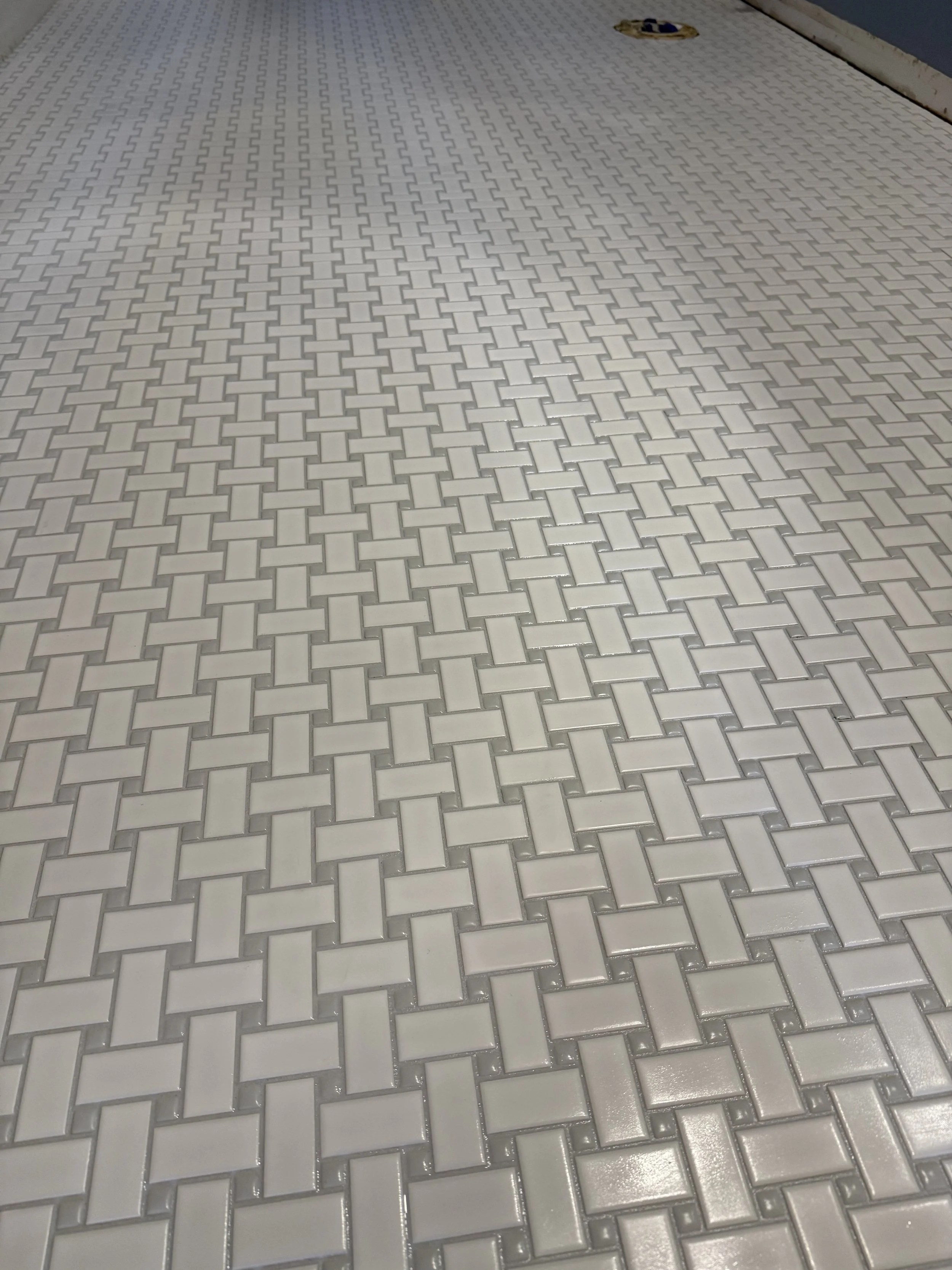 White square tiles arranged in a herringbone pattern on a bathroom floor, with a drain cover visible in the upper right corner.