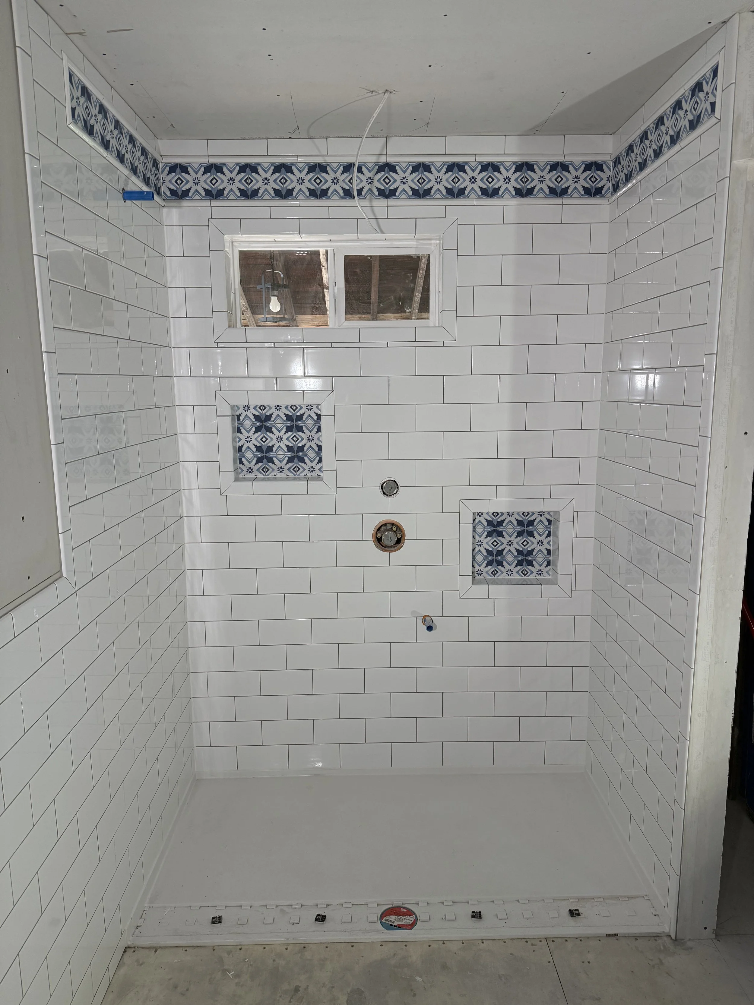 The photo shows a bathroom shower area with white subway tiles on the walls, decorated with a blue and white patterned border tile. There are two recessed shelves with the same patterned tile. A window is visible in the upper center of the wall, and plumbing fixtures are not yet installed. The shower drain and a protective plastic cover are visible at the bottom.