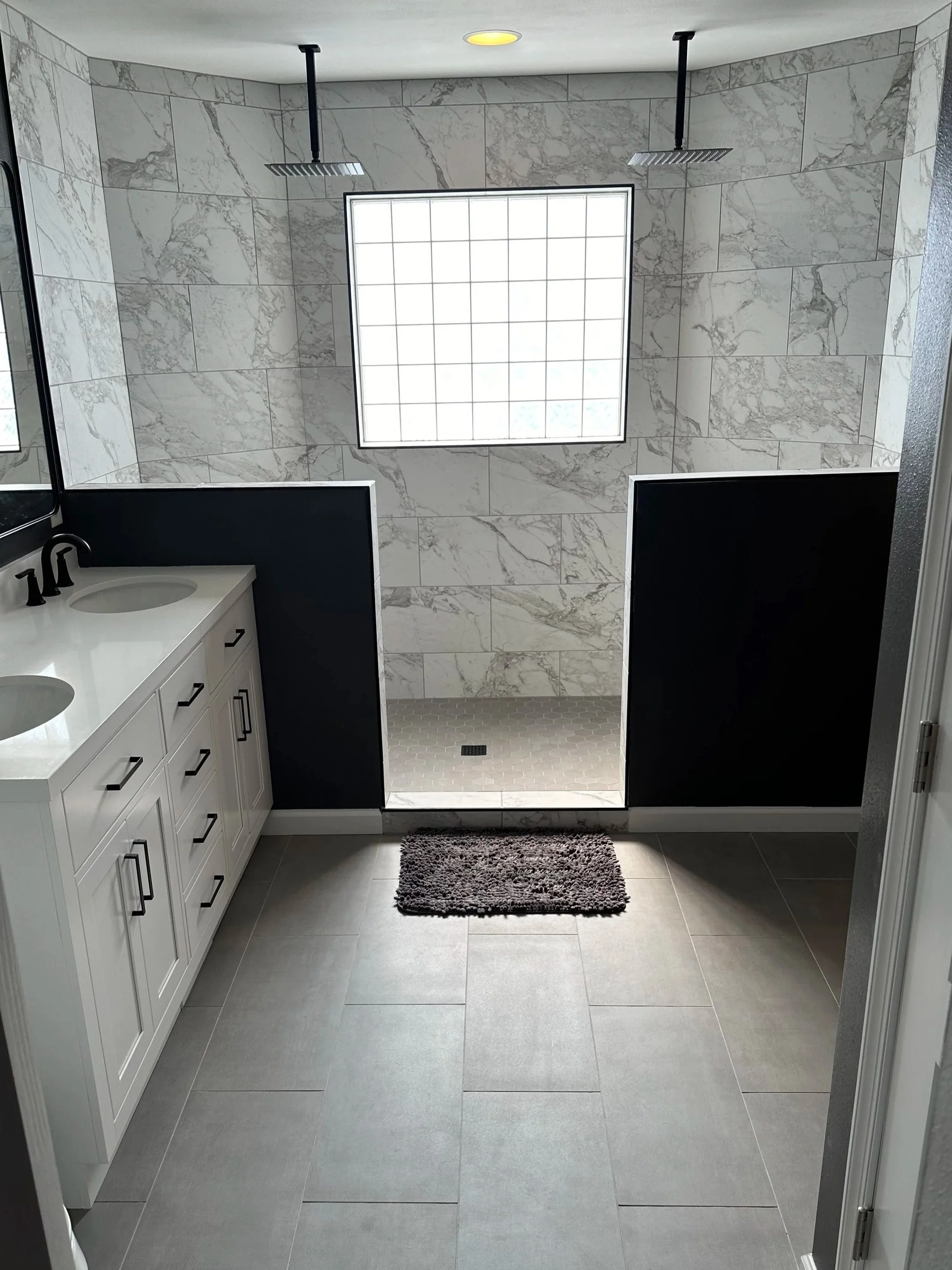 Modern bathroom with large gray tiles, dual shower heads, black accents, a vanity with double sinks, and a window bringing in natural light.