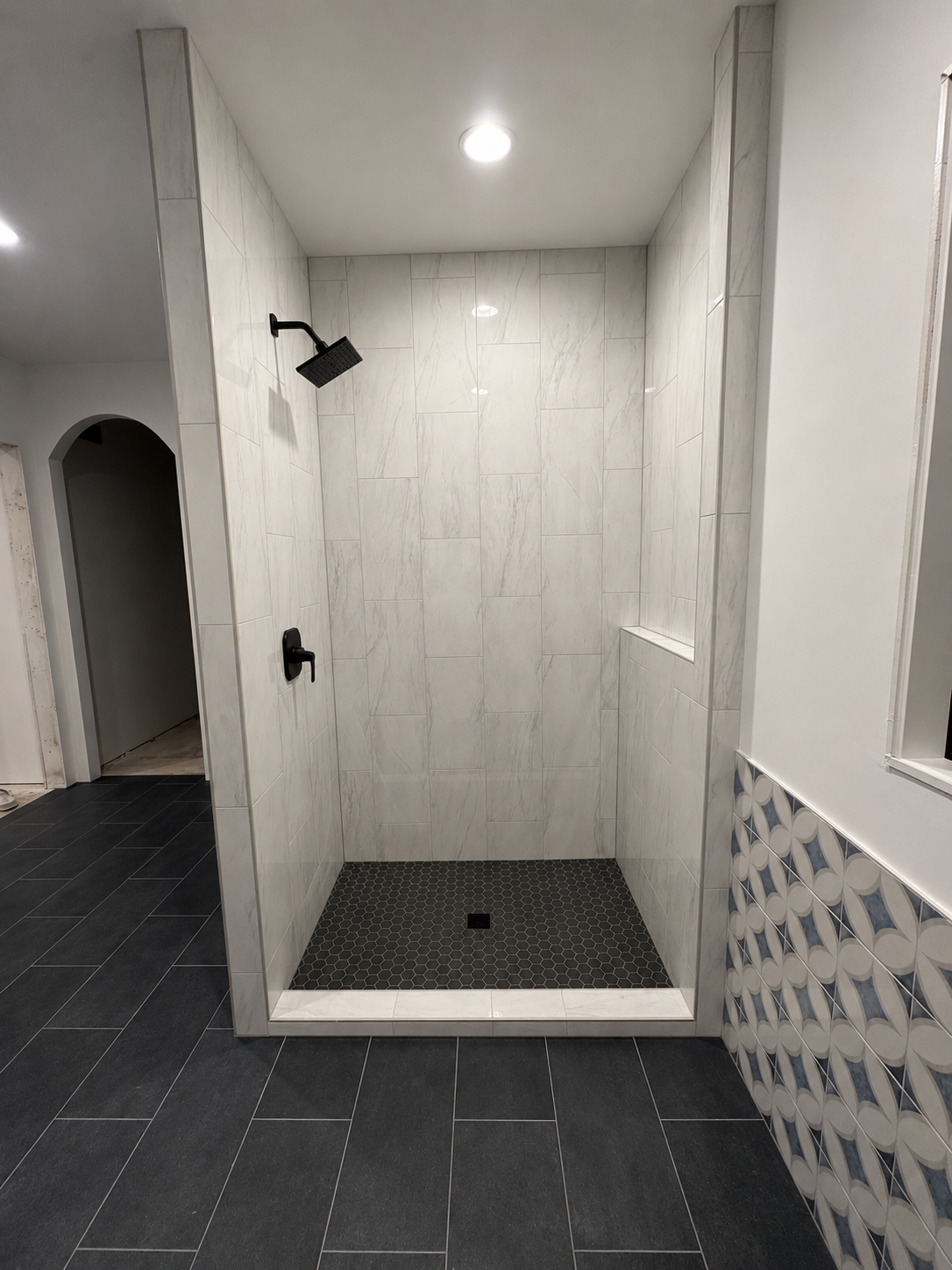 Empty walk-in shower with white marble tiles, black showerhead and faucet, black hexagon tiles on the floor, and a built-in small shelf, surrounded by dark floor tiles and patterned wall.