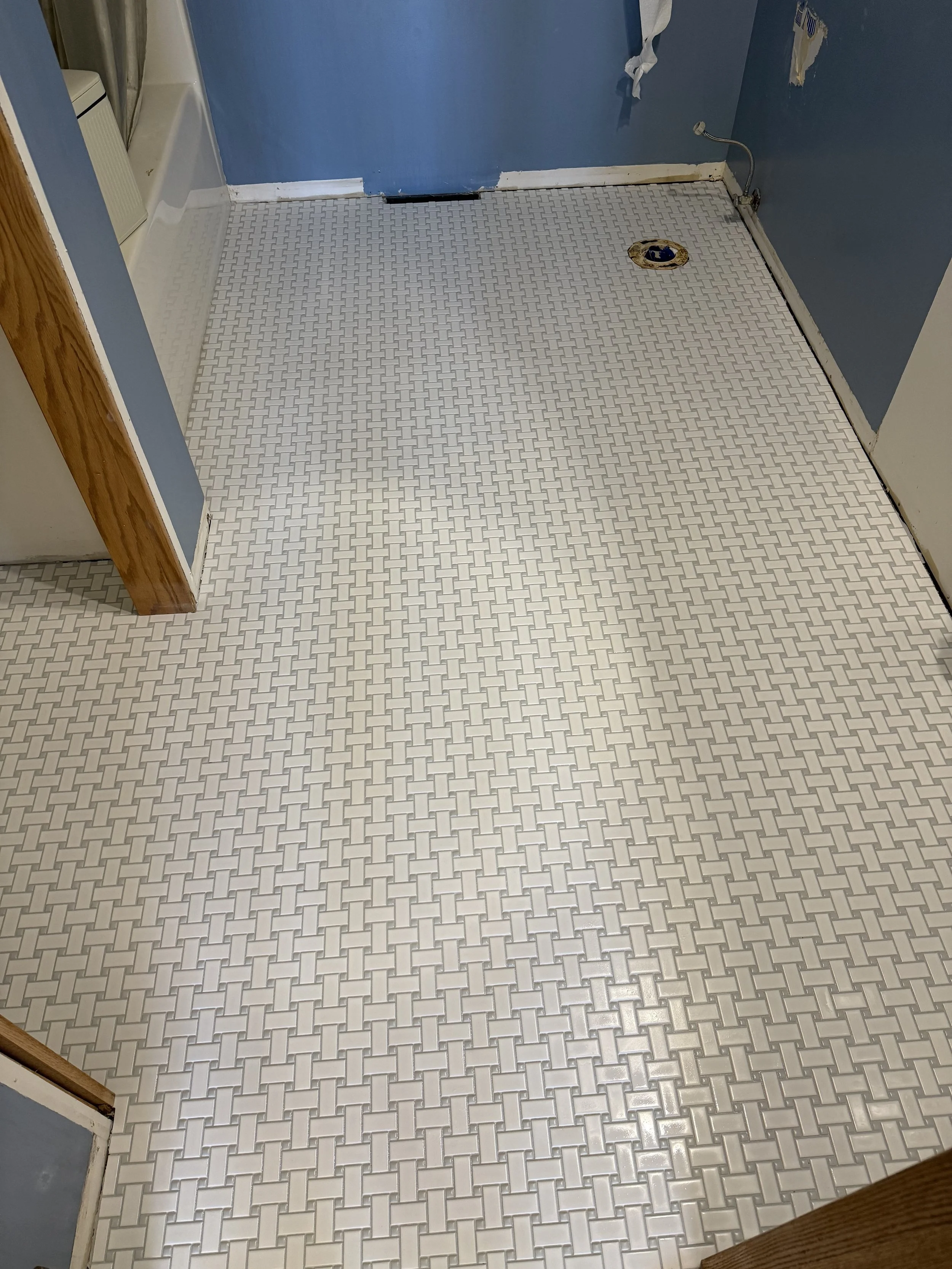 Empty room with white patterned tile floor, blue walls with some damage, a small floor drainage hole, and exposed plumbing.