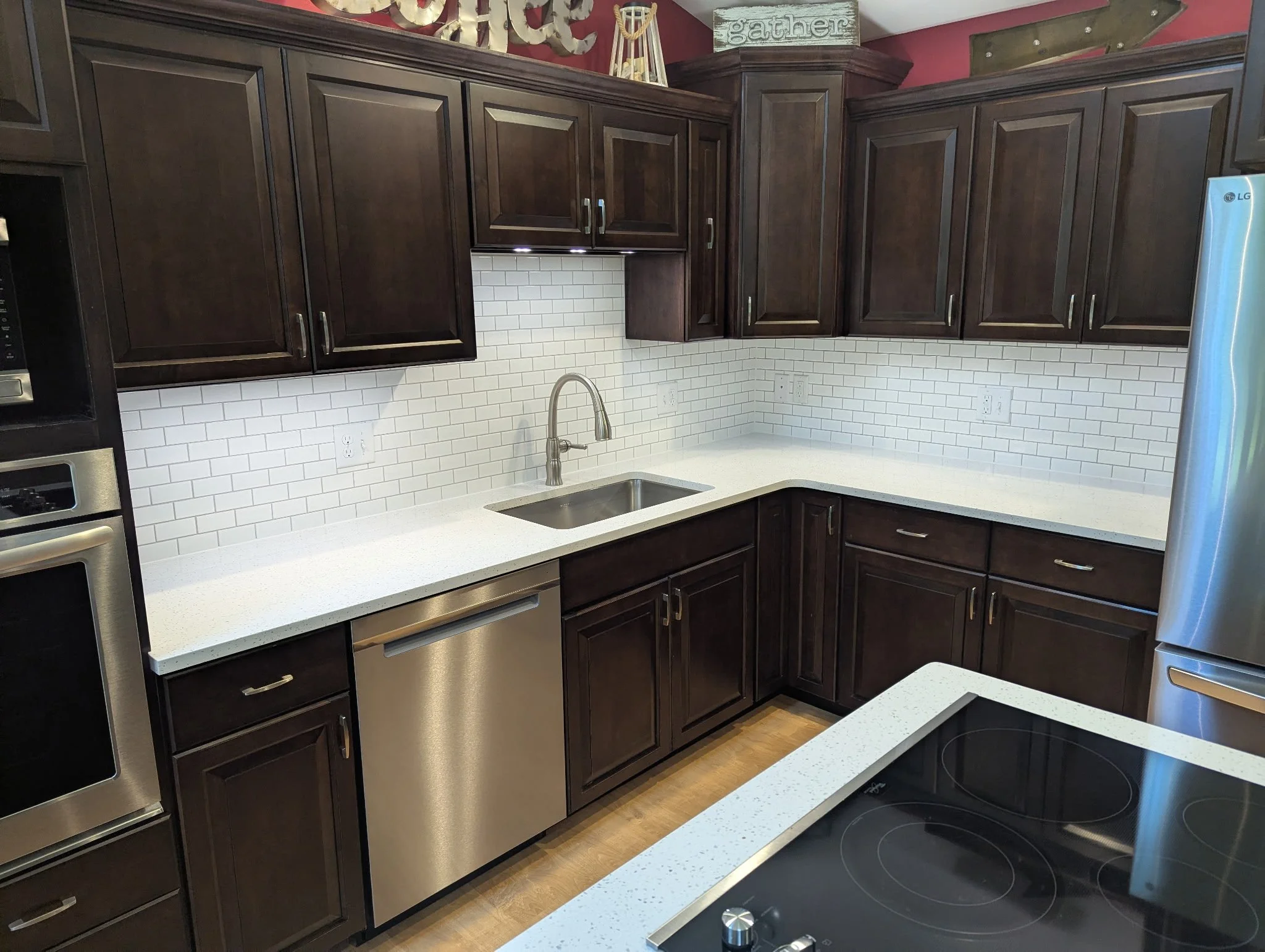 A modern kitchen with dark wood cabinets, a white countertop, a stainless steel sink, and a black electric cooktop. The backsplash is made of white subway tiles, and there is a stainless steel oven on the left. A refrigerator is visible on the right side of the image.
