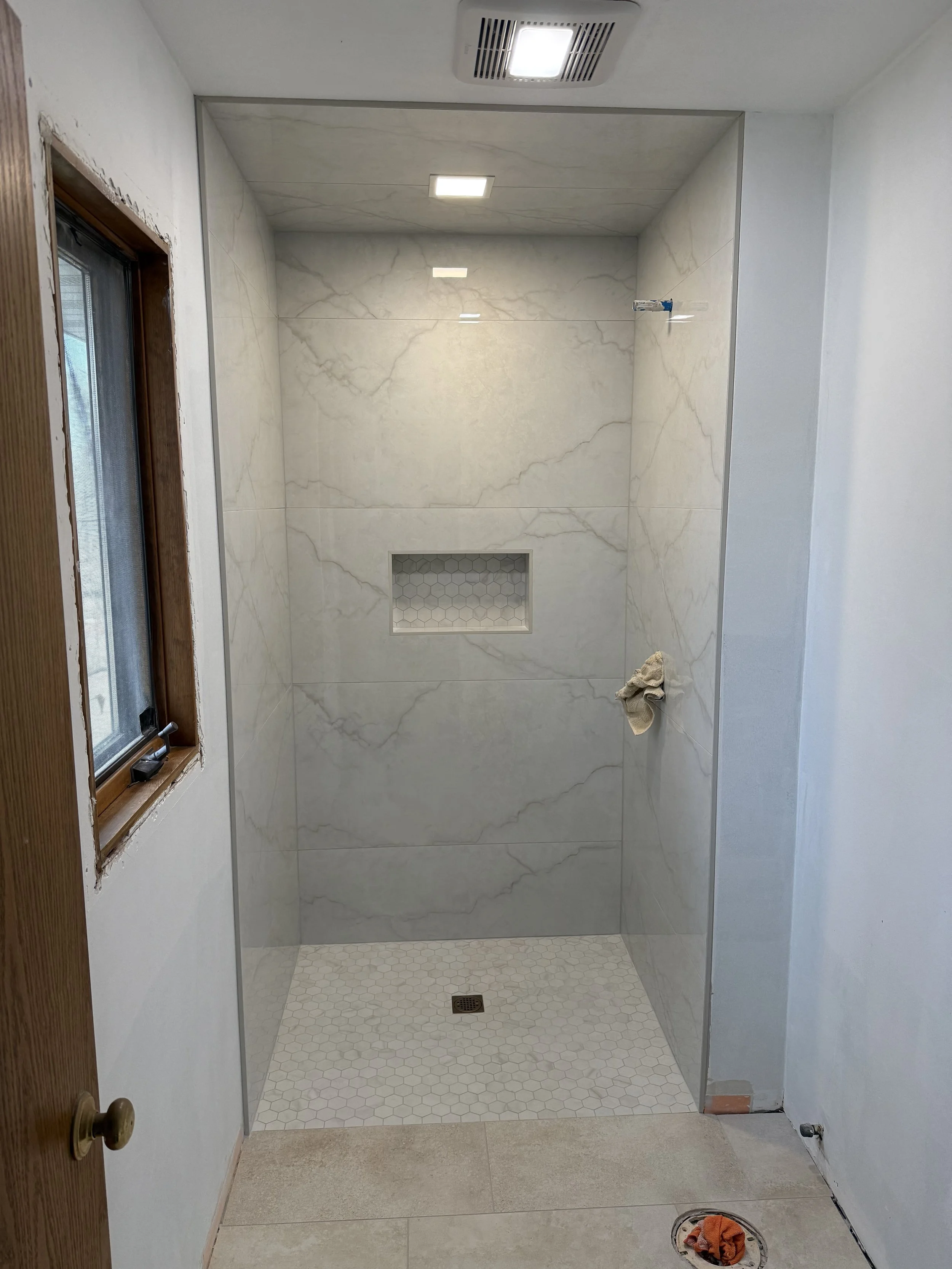 A bathroom shower area with marble walls and a small hexagon-tile floor. There is a built-in niche in the wall with tiles, a drain in the floor, and a towel on the floor near the entrance. A window with a wooden frame is on the left side, and there are ceiling lights and an exhaust vent.