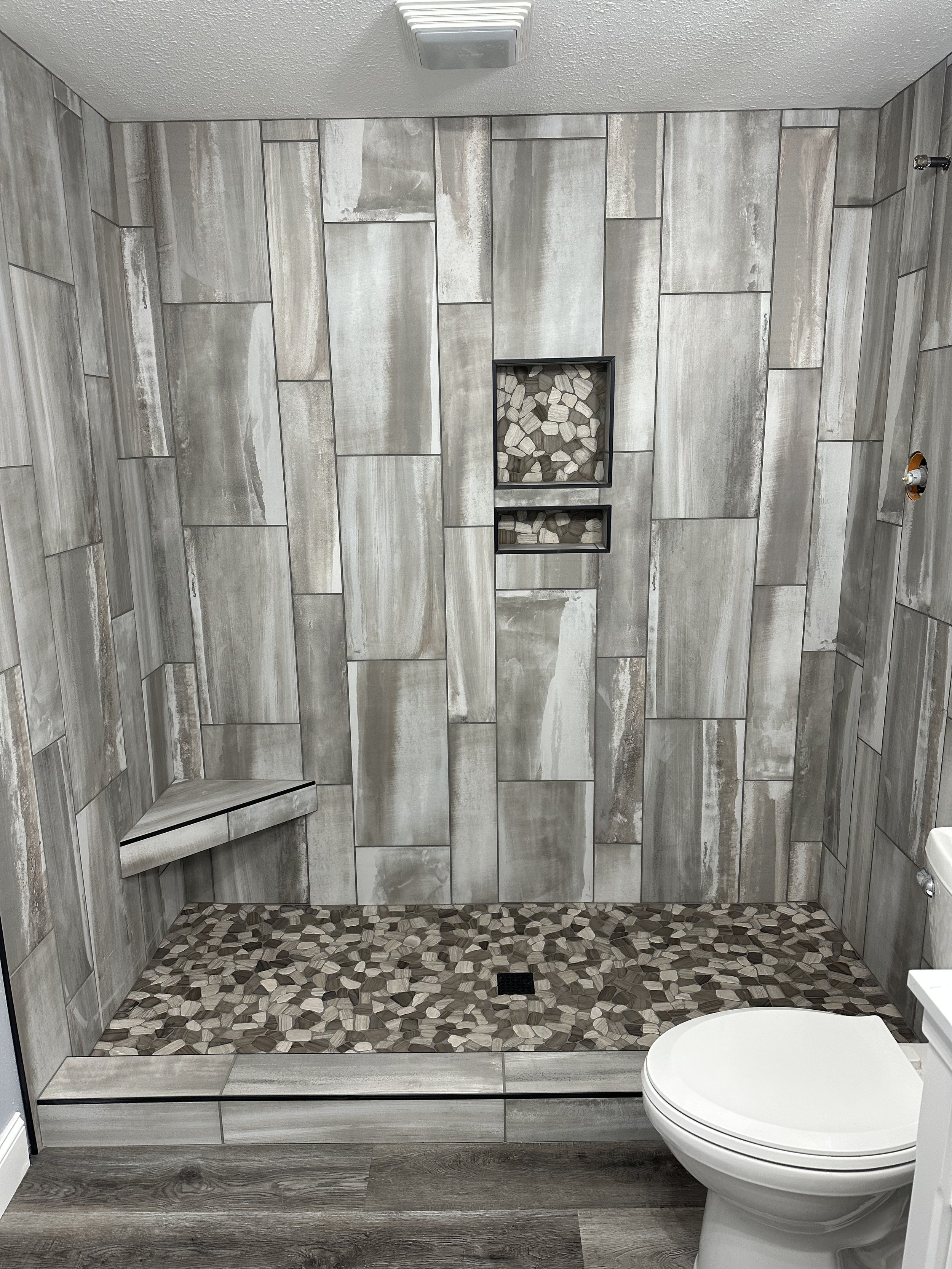 Modern bathroom with tiled shower area that has a built-in bench, marble-like wall tiles, and pebble floor tiles. A toilet is visible in the foreground.