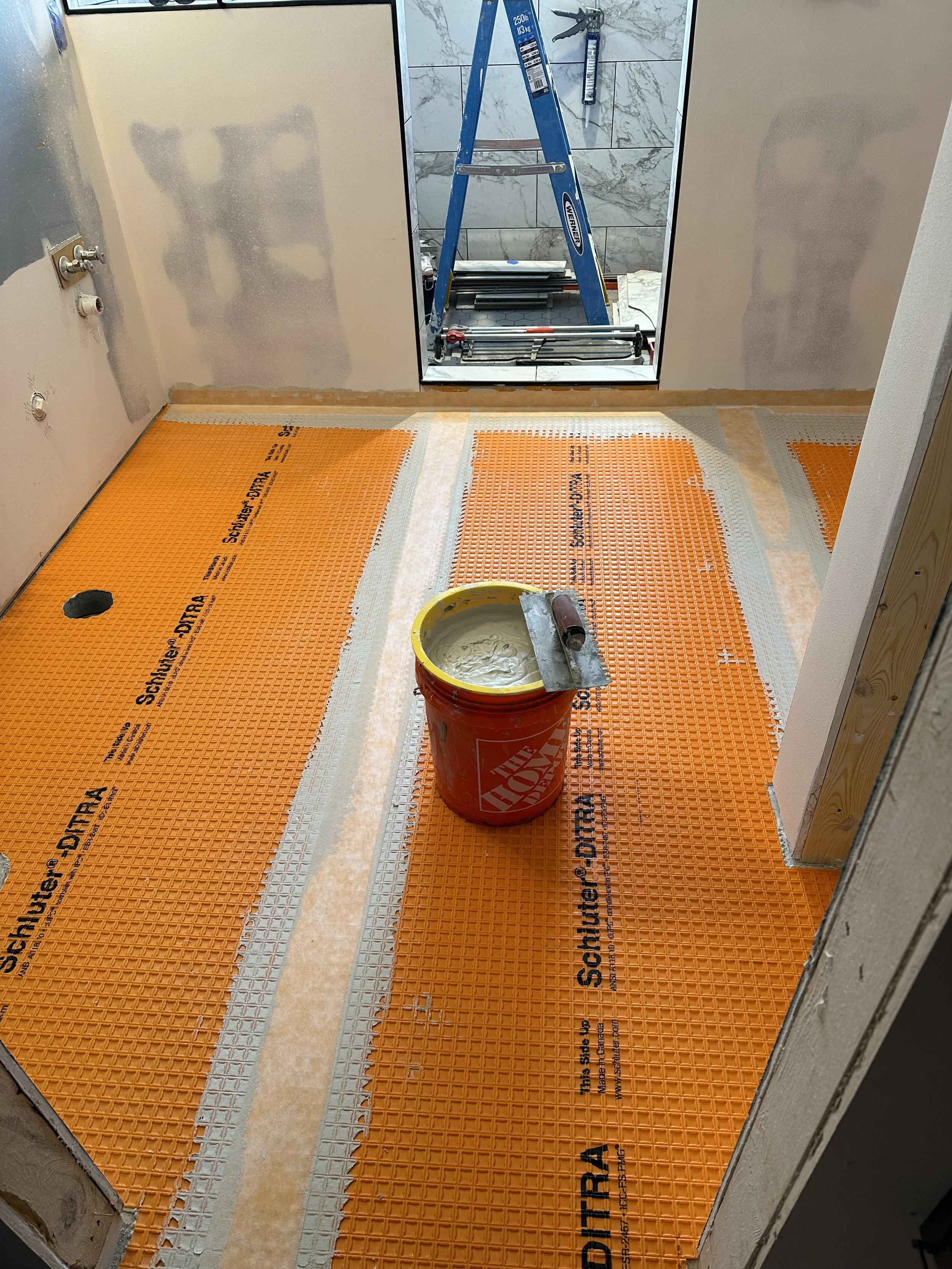 A bathroom under construction with orange Schluter shower membrane on the floor, a bucket of waterproofing or sealant, a trowel, and a blue ladder in front of a mirror. Part of the wall shows drywall with patches of joint compound.