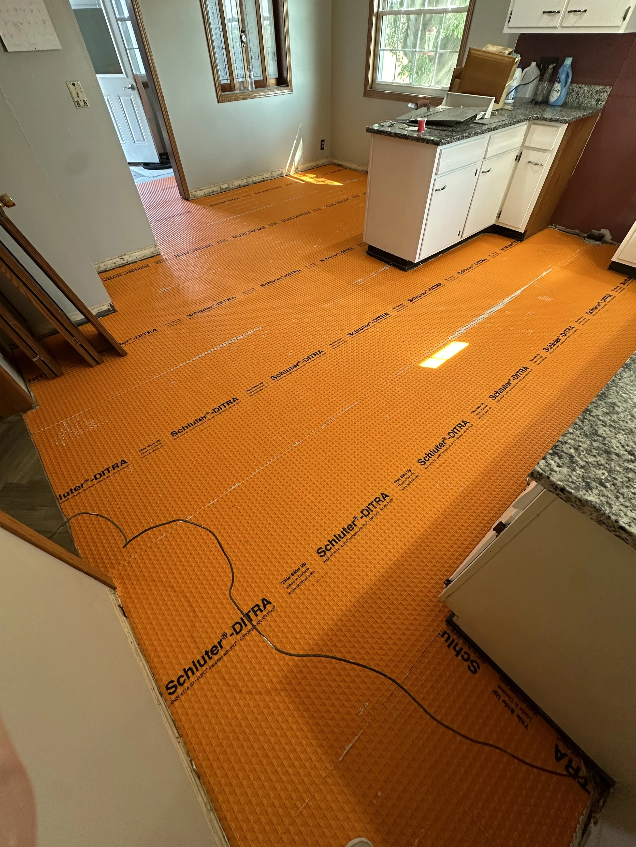 Kitchen with orange Schluter-Ditra flooring material installed. Cabinets and countertops are visible, with some cleaning supplies on the counter. A window and a door leading outside are in the background.