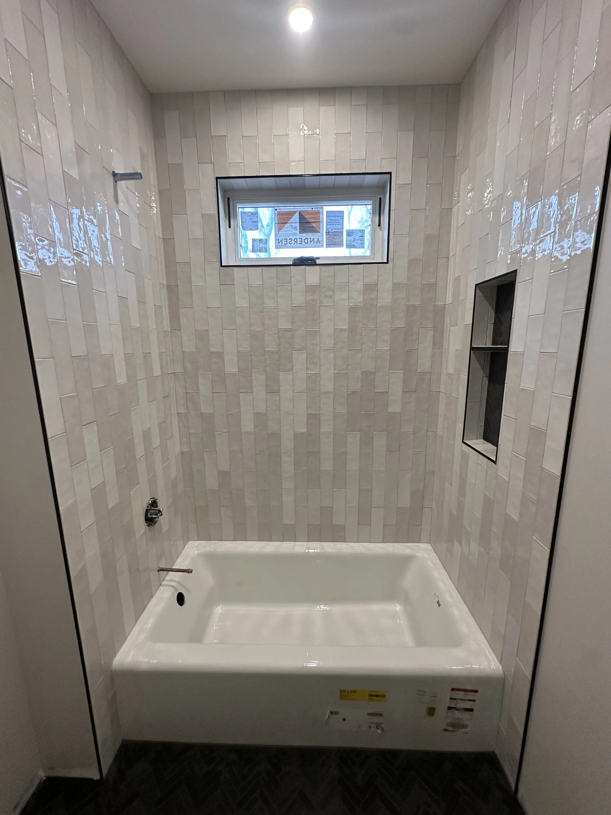 An empty bathroom shower stall with beige patterned tiles on the walls, a small window, and built-in shelving.