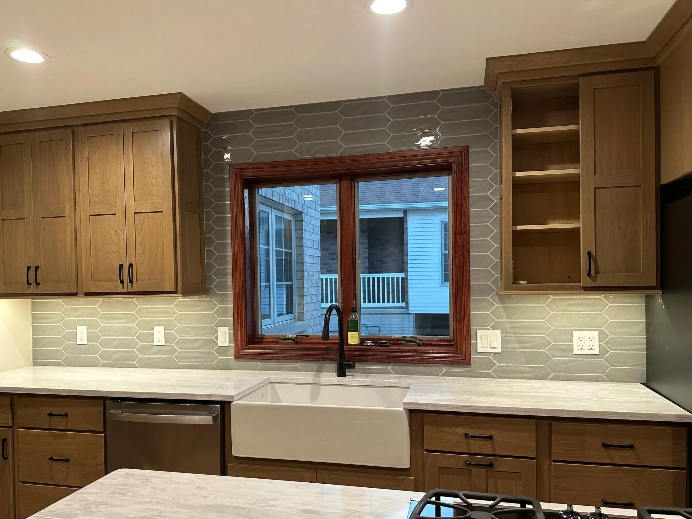 Kitchen with wooden cabinets, gray hexagonal tile backsplash, a window showing an outdoor patio, white countertops, and a black faucet.