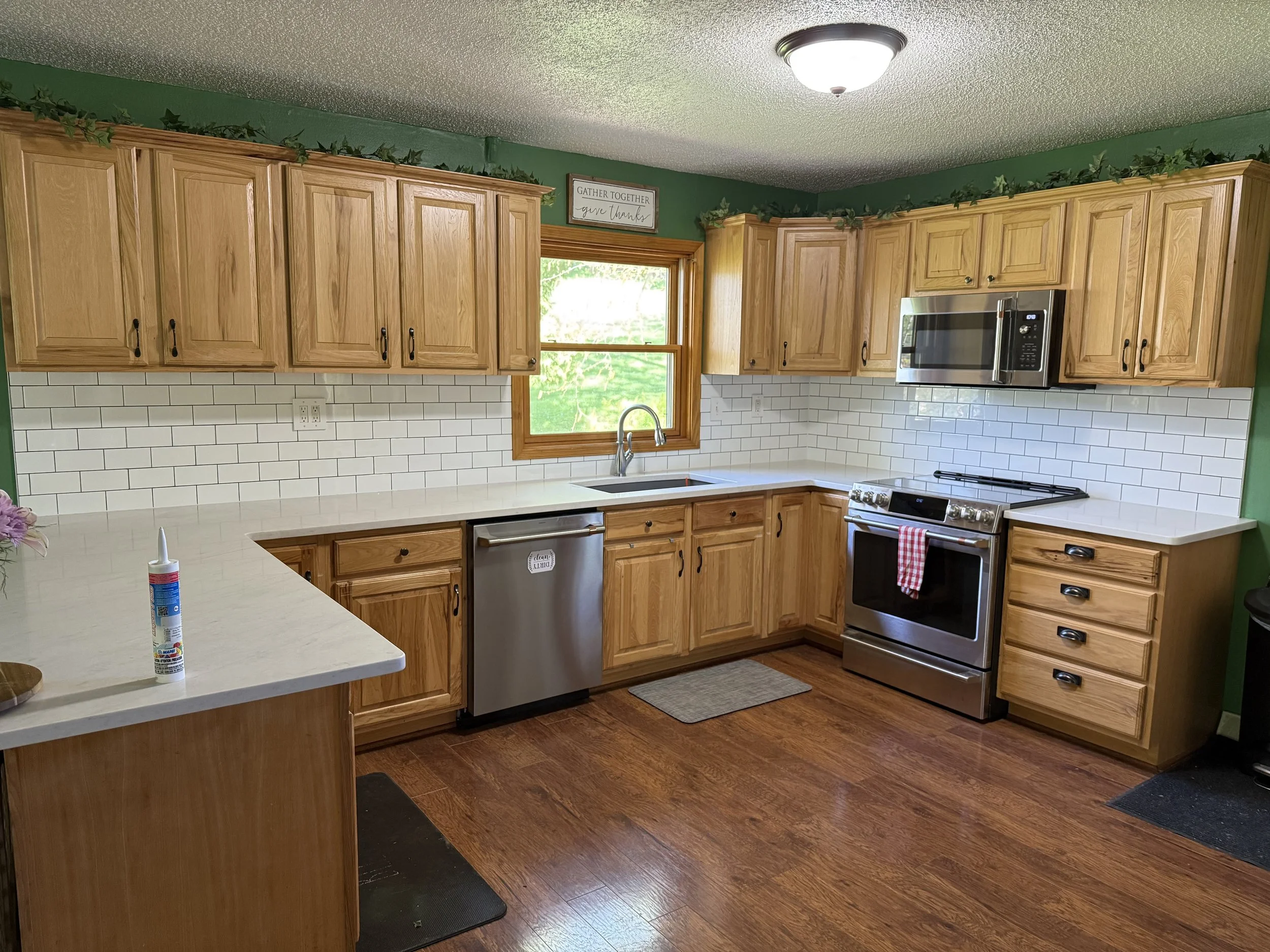 Kitchen with wooden cabinets, white subway tile backsplash, stainless steel appliances, a window above the sink, and a ceiling light fixture. Green walls and decorative greenery above the cabinets.