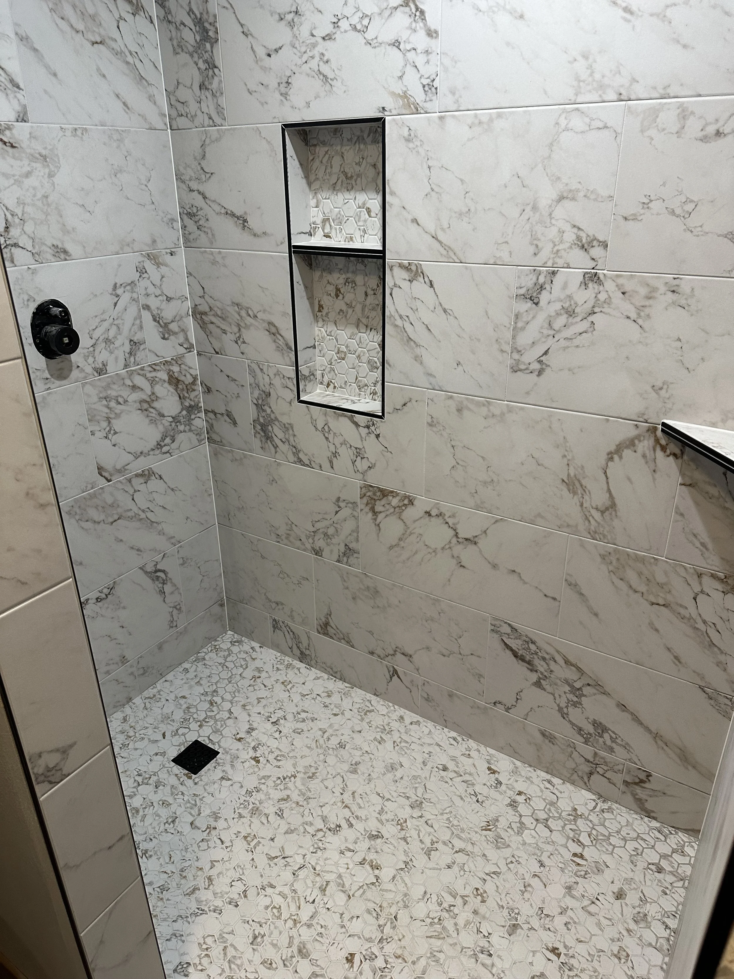 A modern walk-in shower with large marble-looking tiles on the walls and a small hexagon tile pattern on the floor. It has two built-in wall niches with honeycomb-shaped tiles, a black drain on the floor, and a black fixture with a trigger on the left wall.