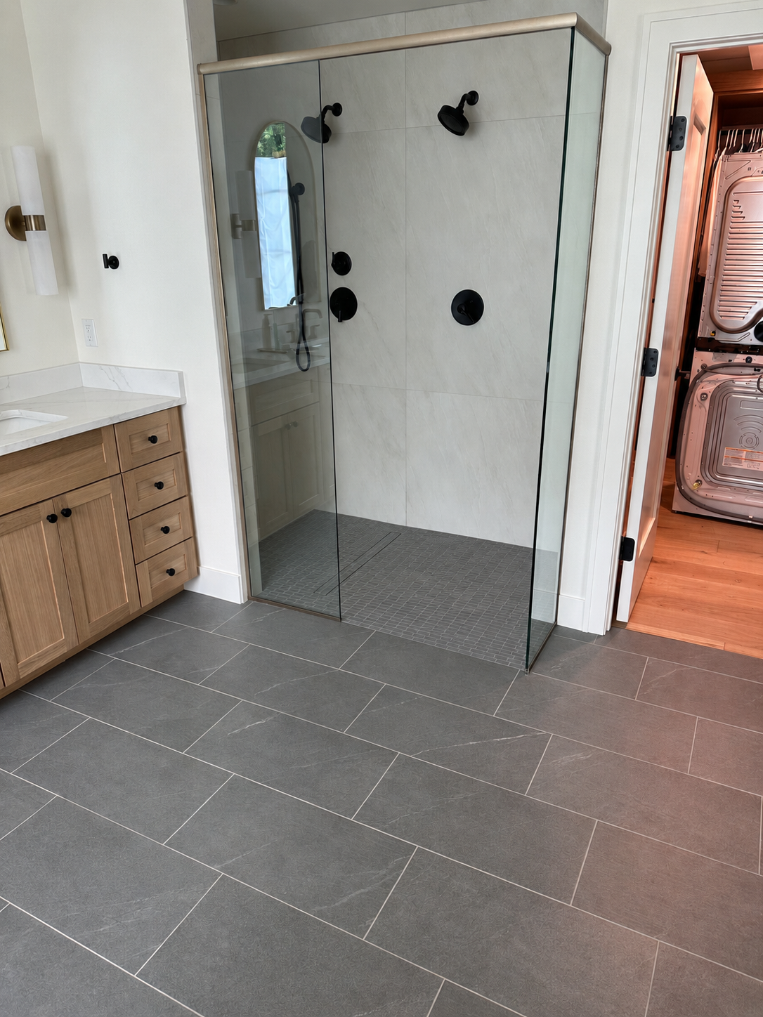 A modern bathroom with a glass shower enclosure, black fixtures, a wooden vanity with a white marble countertop, and a laundry room with stacked washer and dryer visible through an open door.