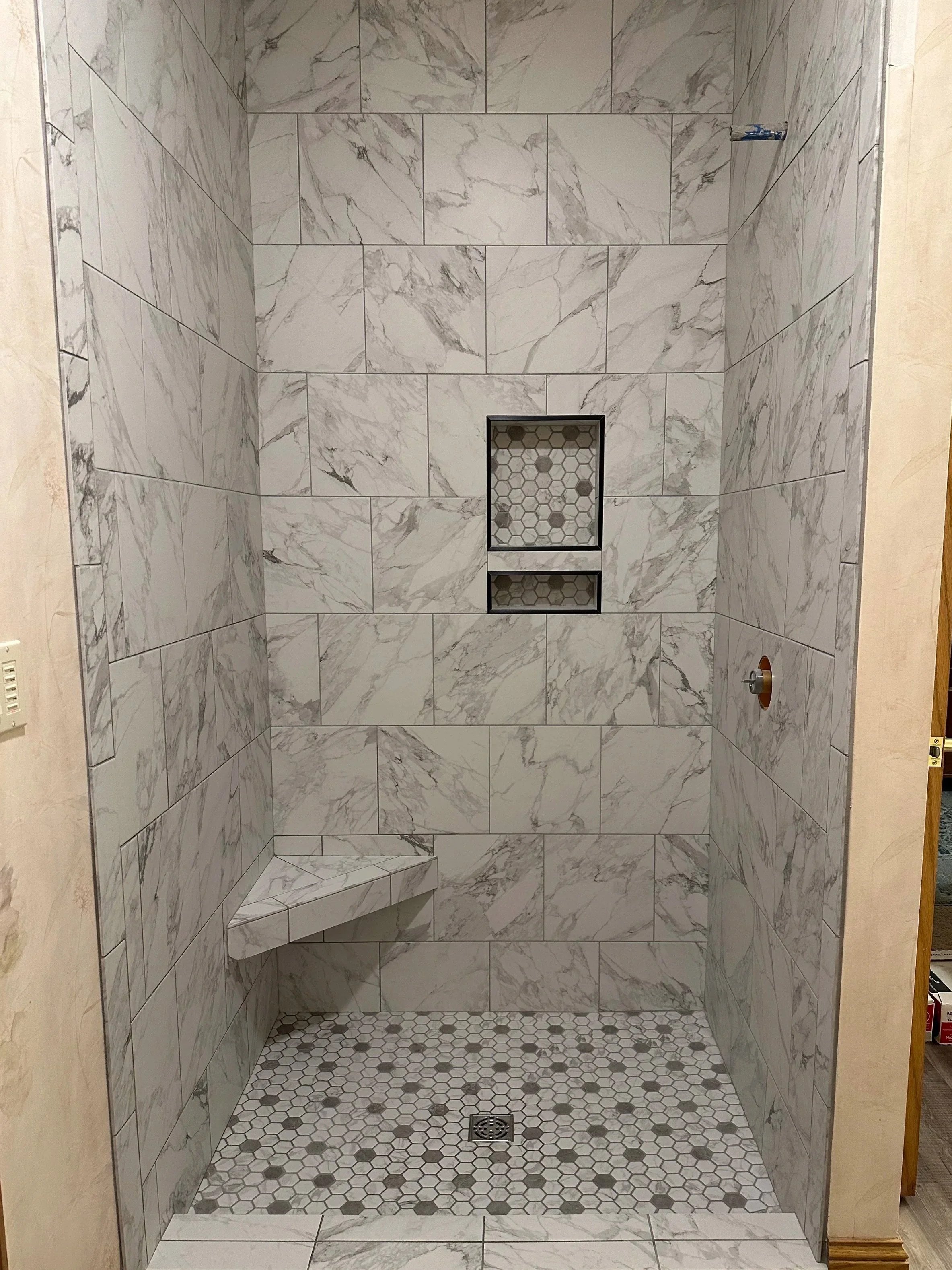 A walk-in shower with marble tiles on the walls, small hexagon tiles on the floor, a built-in corner bench, and two niche shelves with hexagon tile backing in the center.