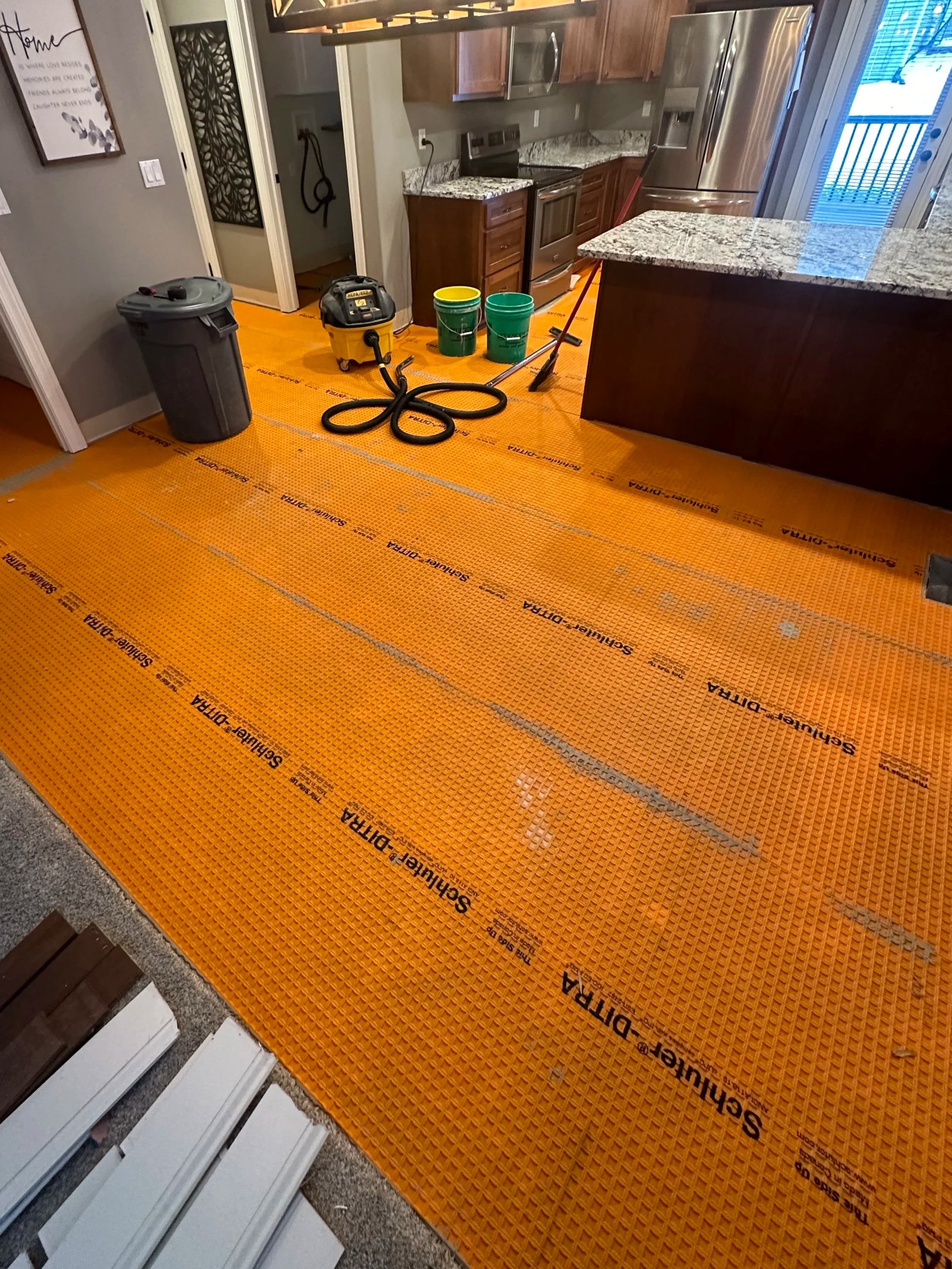 Kitchen with orange safeguard underlayment on the floor, countertops, stainless steel refrigerator, oven, microwave, and various cleaning tools including a vacuum, mop, and buckets.