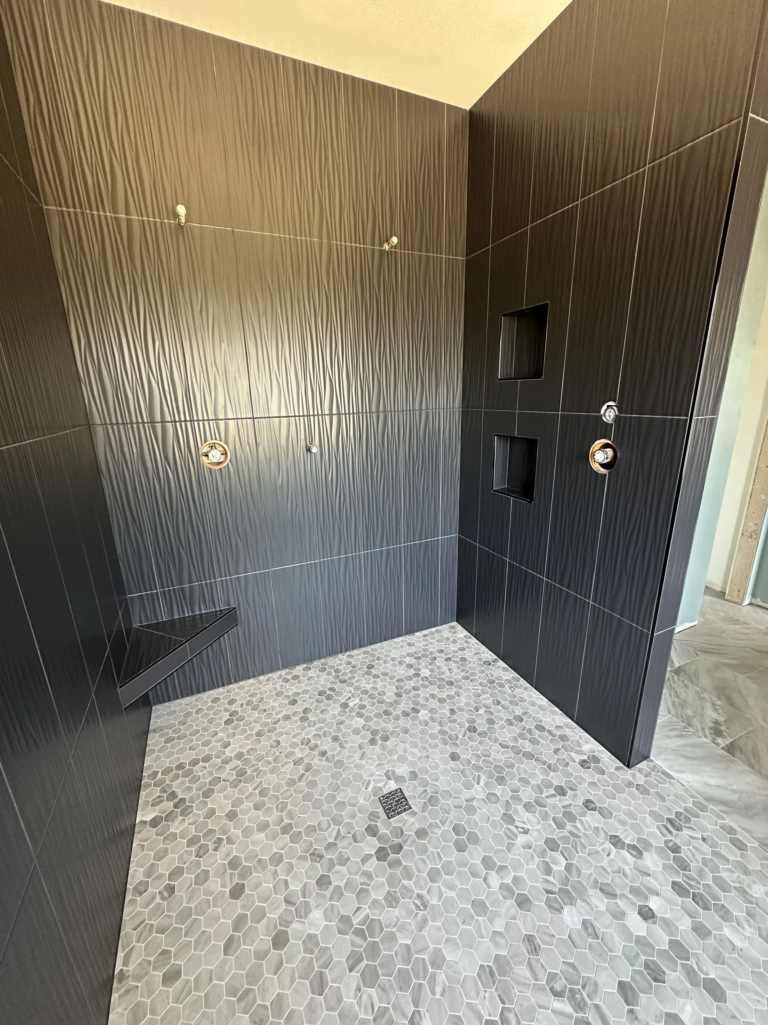 Modern shower area with dark tile walls, small round hexagonal floor tiles, a built-in corner bench, two niche shelves, and exposed plumbing fixtures.
