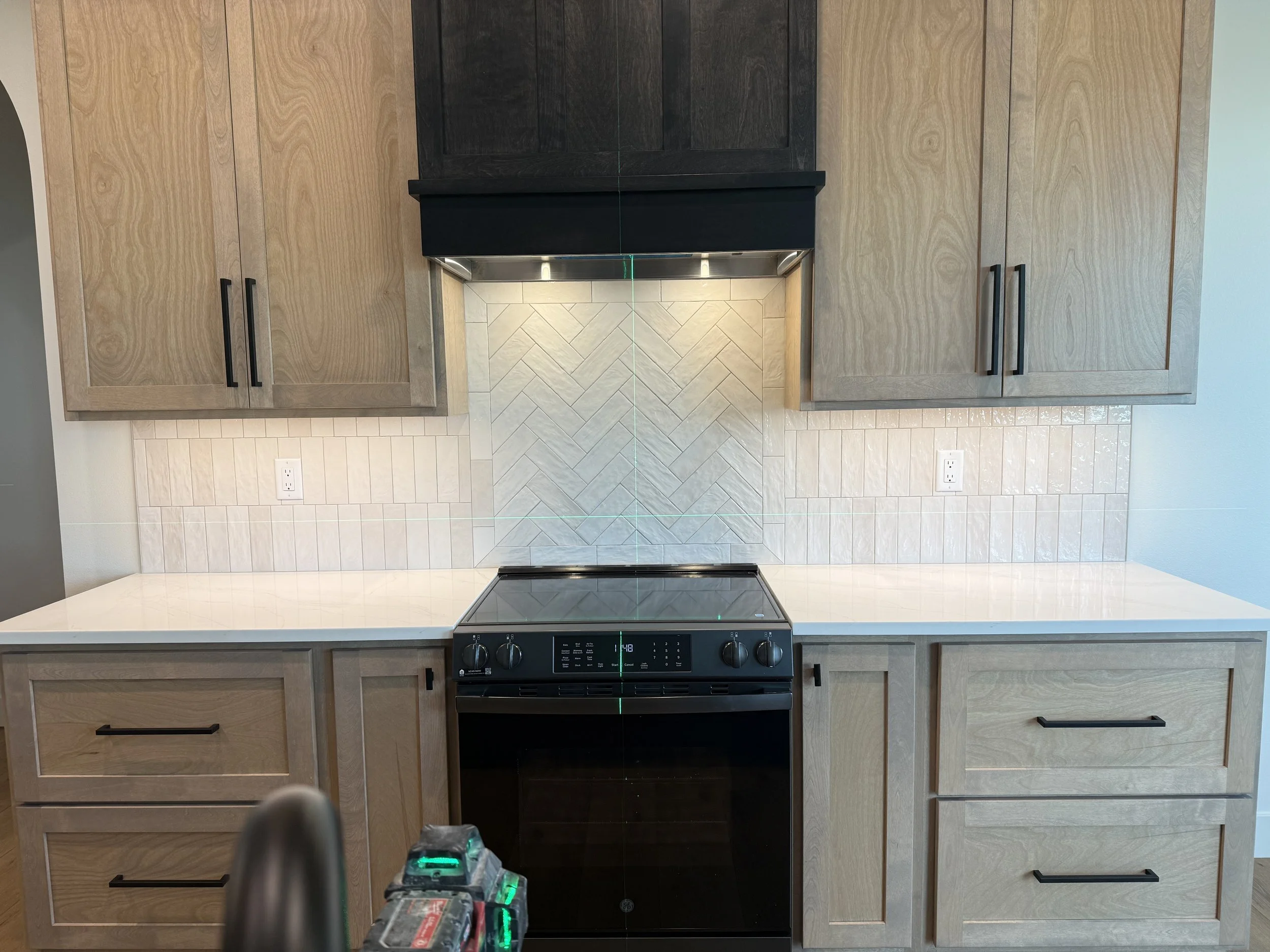 Kitchen with light wooden cabinets, a black range hood, a black stove, a tiled backsplash, and power outlets on the wall.