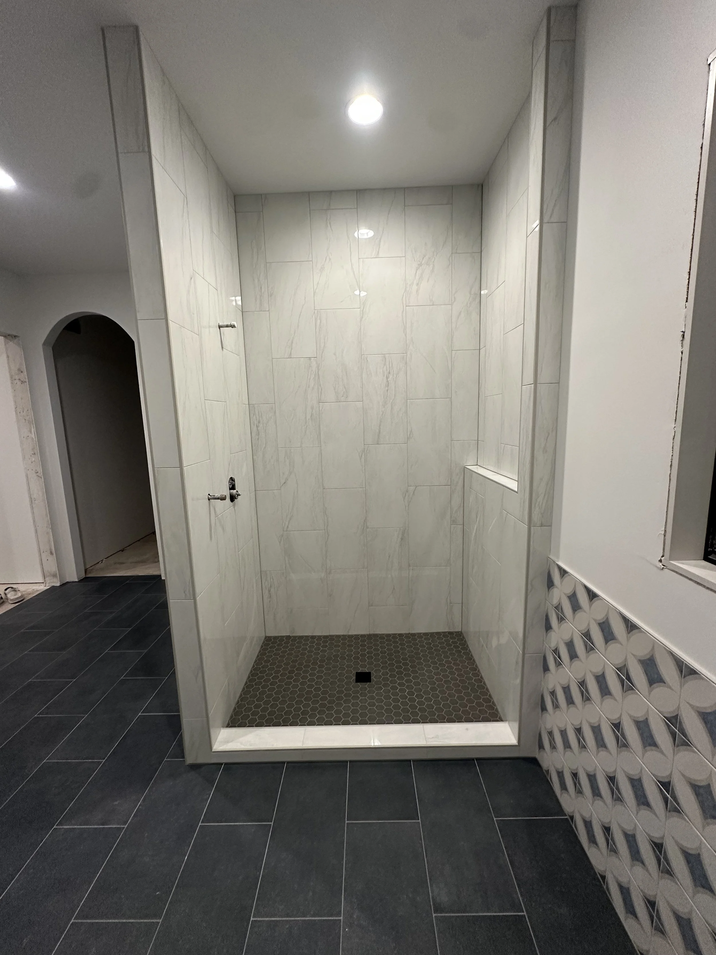 An unfinished bathroom shower with marble tile walls and a small recessed niche. The shower floor has small hexagonal tiles, and the shower is framed by white walls with a patterned tile accent. The bathroom has dark gray floor tiles.