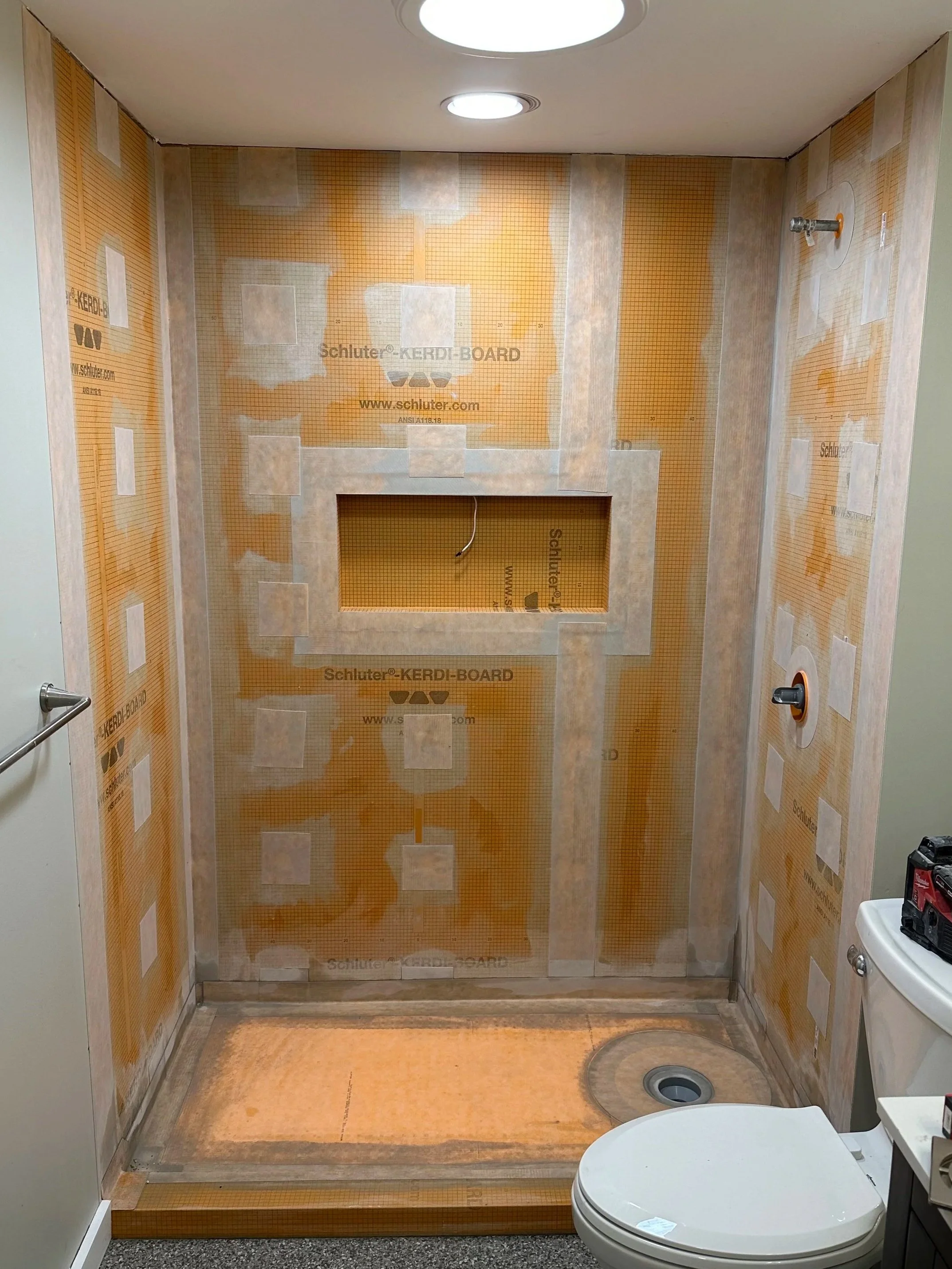 Bathroom under construction with unfinished shower area, drywall with insulation behind plastic sheeting, a toilet in the foreground, and construction materials.