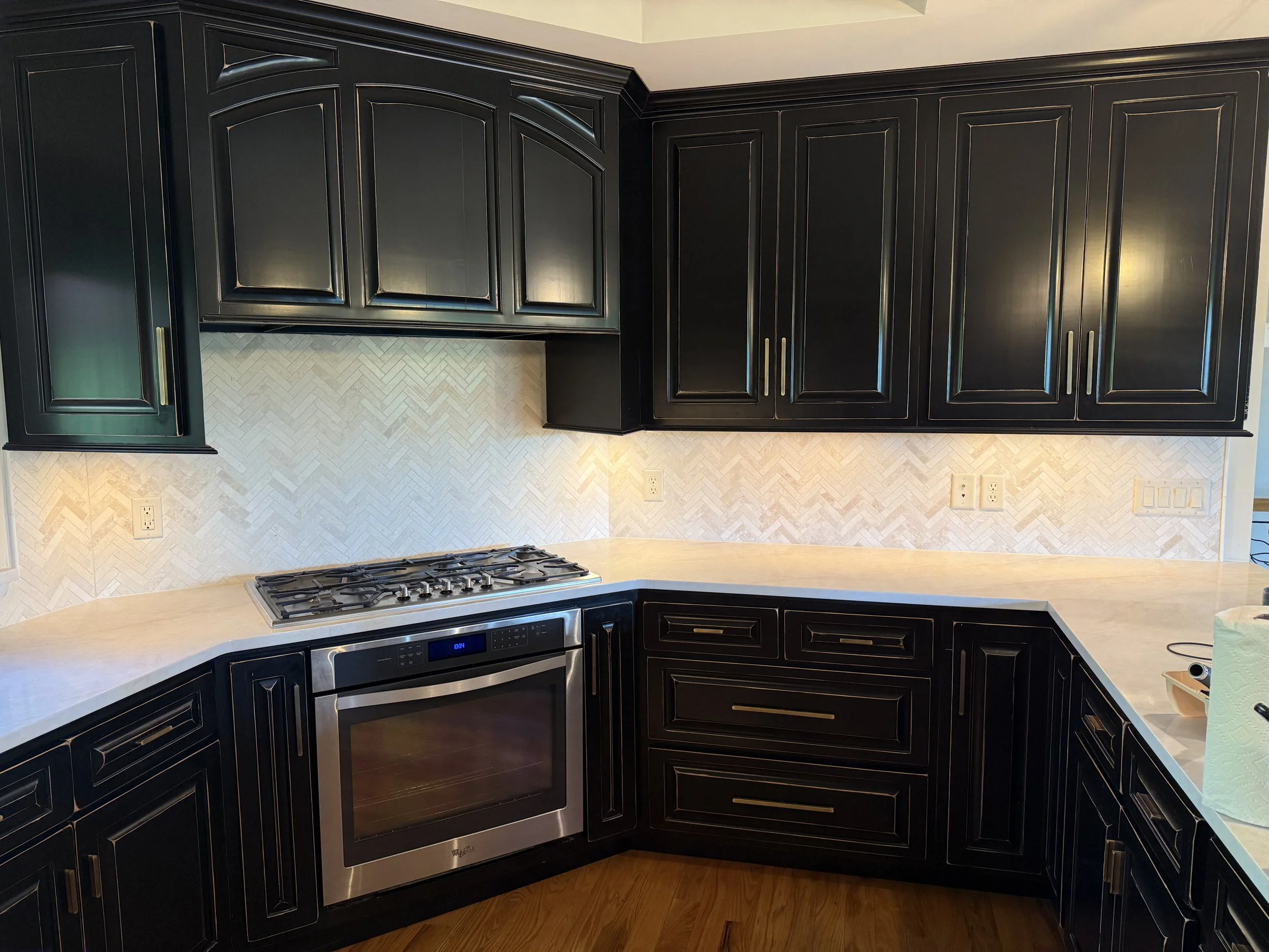 Black kitchen cabinets with gold handles, beige herringbone tile backsplash, stainless steel stove, and beige countertop.
