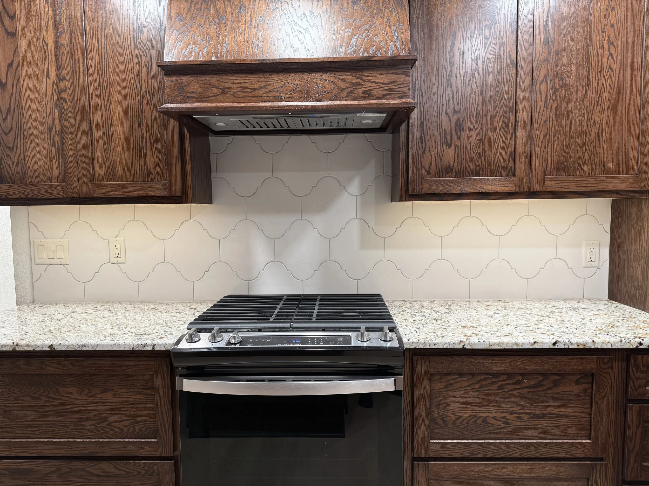 View of a kitchen stovetop with a granite countertop and dark wood cabinets, white tile backsplash, and a stainless steel oven.