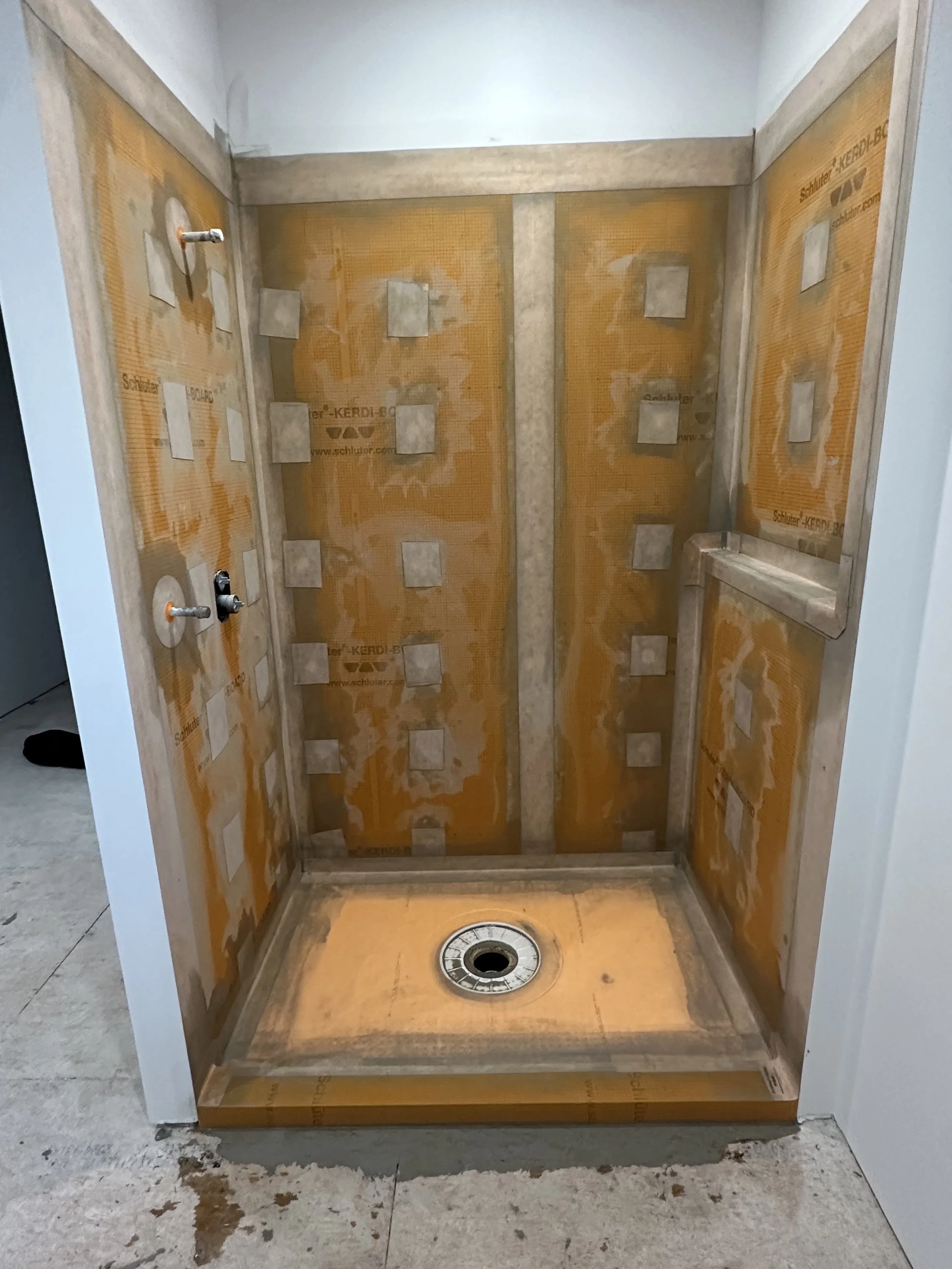 Interior of an unfinished shower with orange waterproofing membrane, wall niches, and a drain at the floor. The construction appears incomplete.