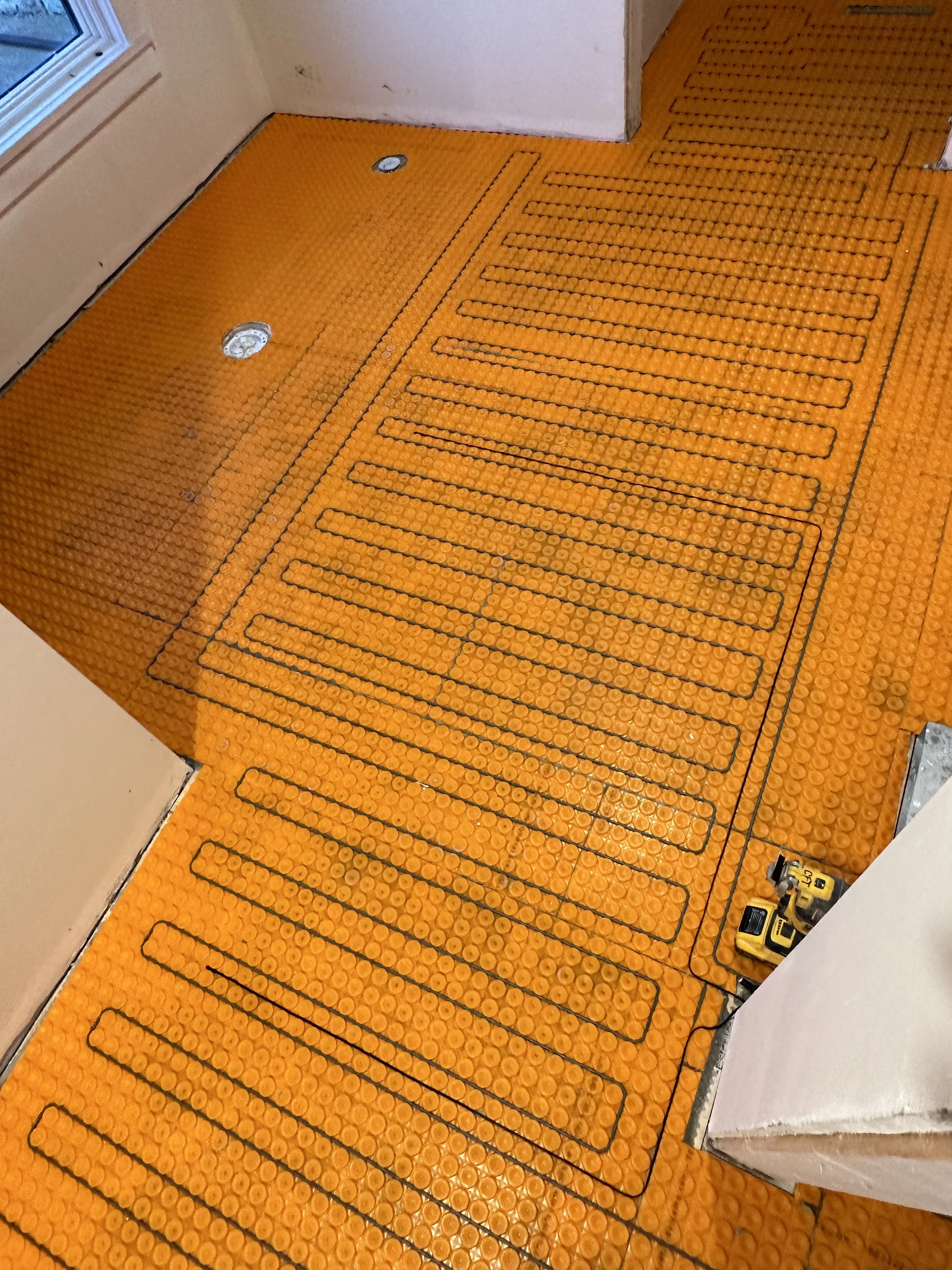 Orange safety flooring with black lines for heating or cable installation, partially installed in a room with unfinished wall and a window.