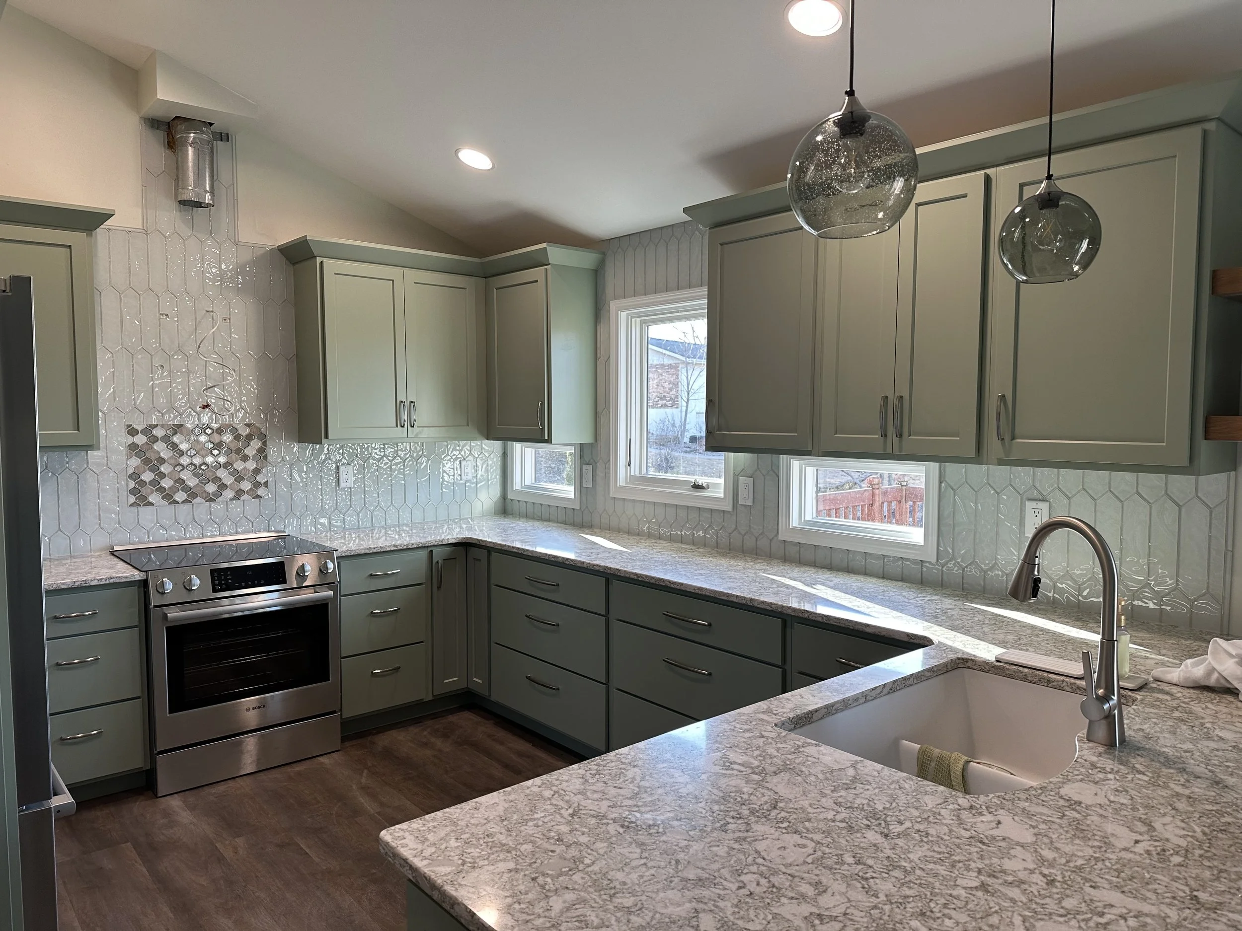 Modern kitchen with green cabinets, white hexagonal tile backsplash, granite countertops, stainless steel oven, and pendant lights.