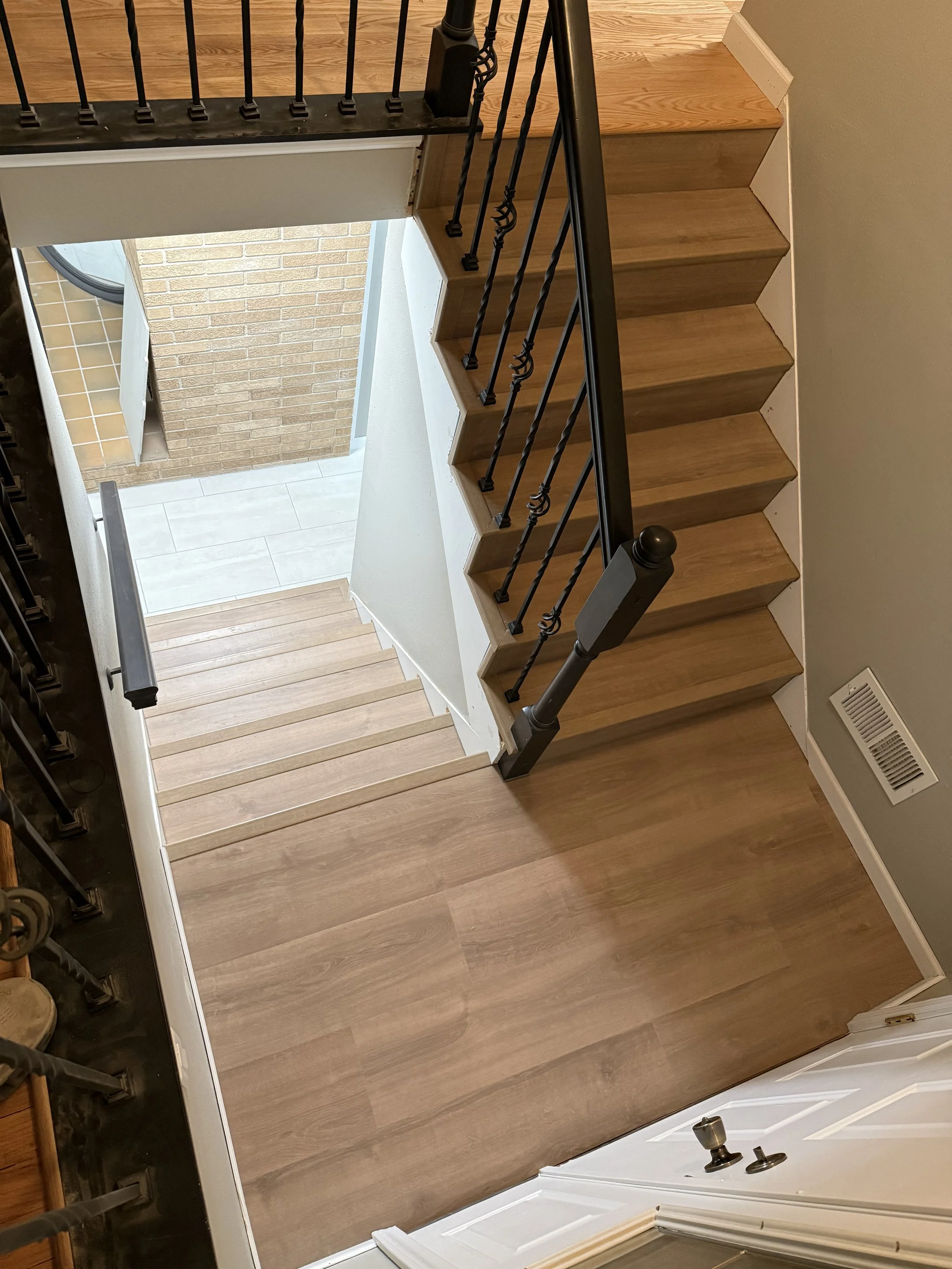 View of a staircase with a wooden floor and a railing made of dark metal spindles, leading down to a tiled entryway with a brick wall and a door.