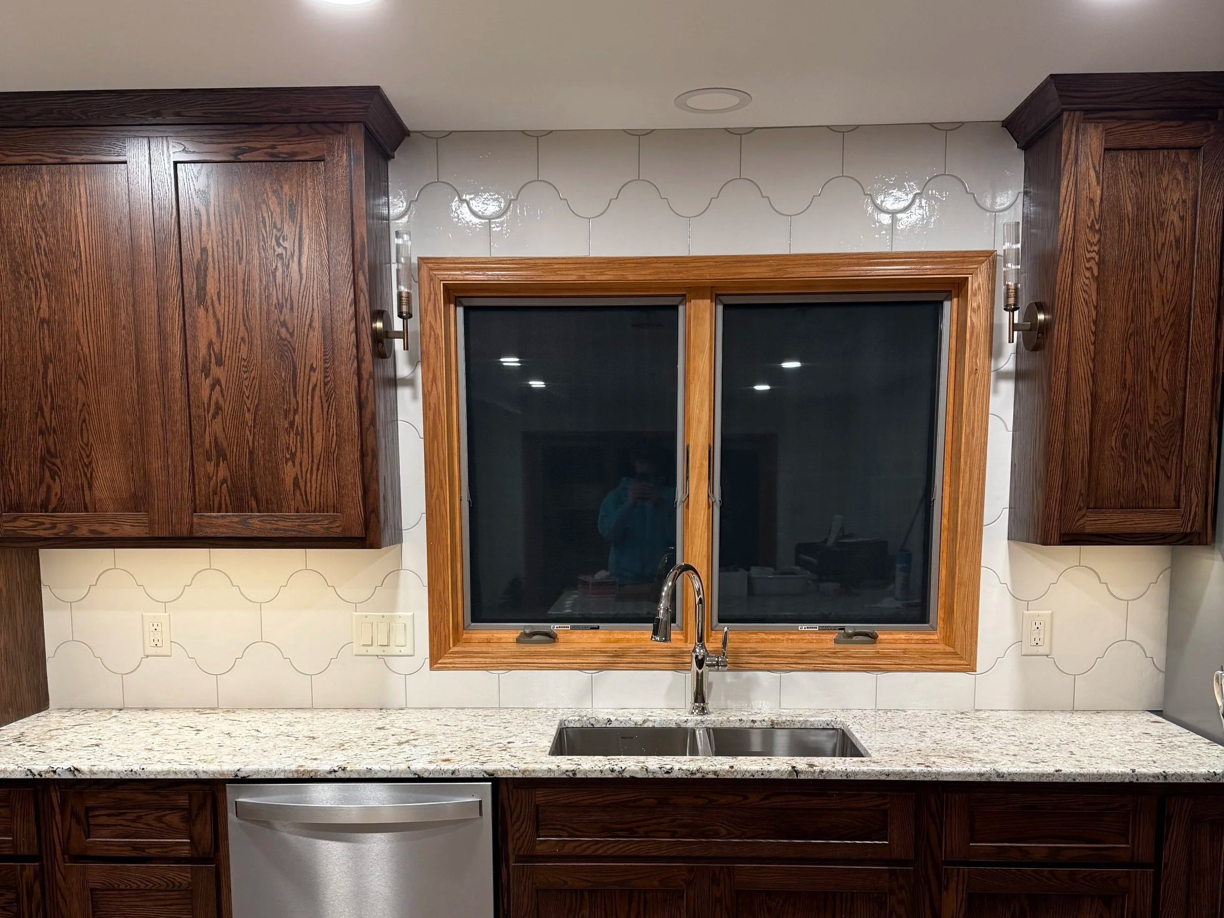 Kitchen with wooden cabinets, a granite countertop, a double-pane window with wooden trim, and a white backsplash with a subtle pattern. There are electrical outlets and a kitchen faucet over the sink.