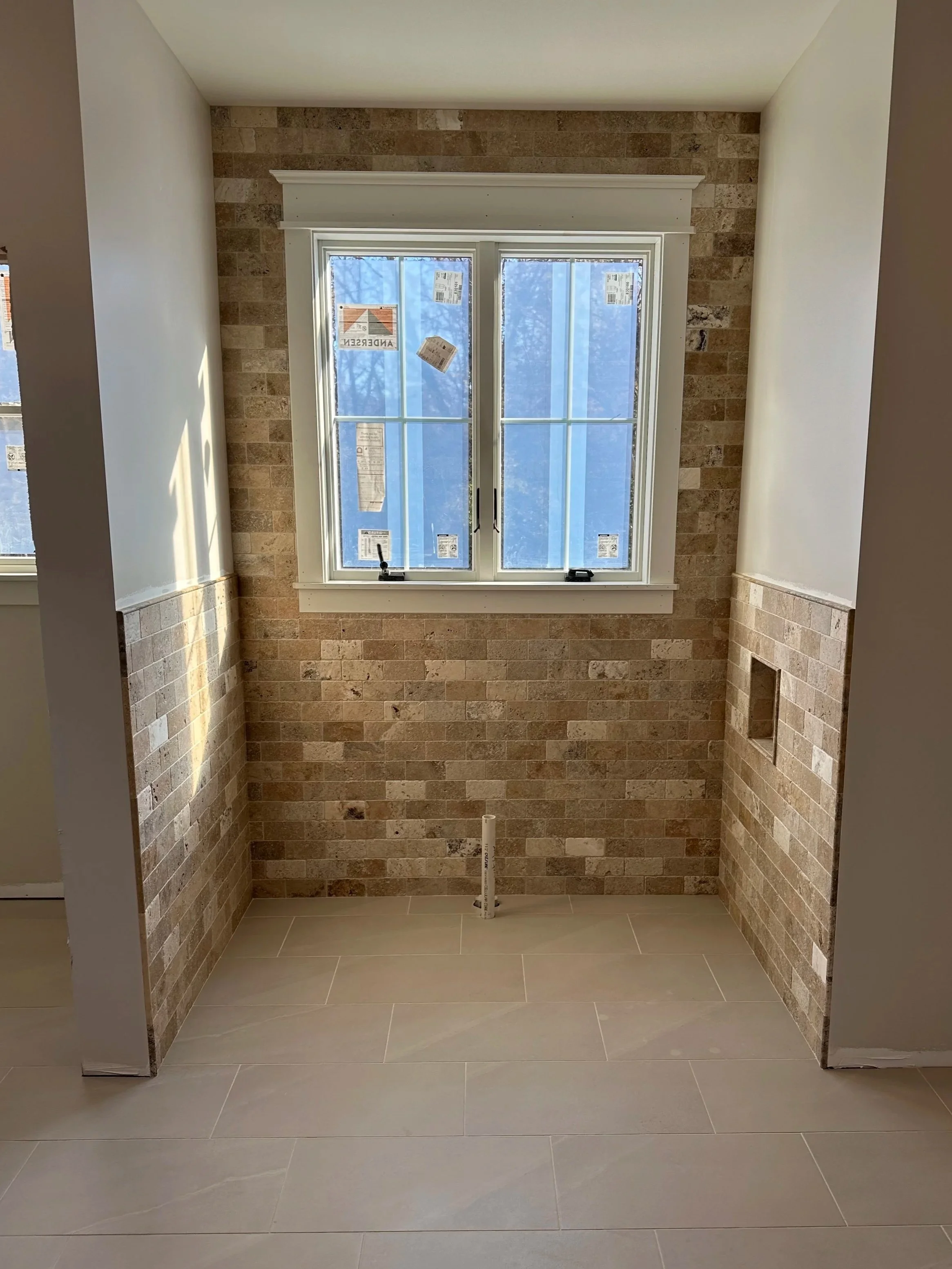 A room under construction with a large window, brick walls, beige floor tiles, and unfinished walls.