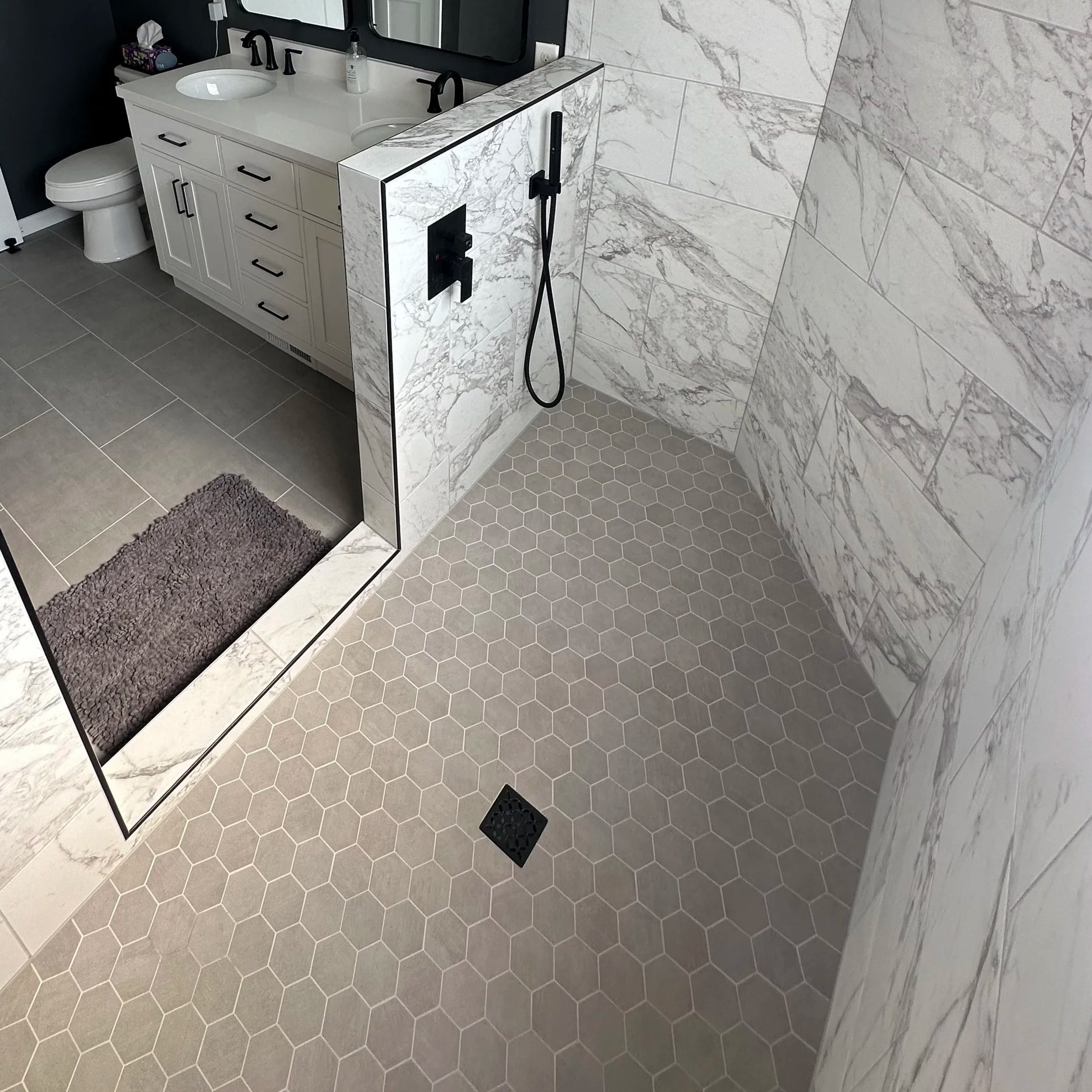 Bathroom with marble tiles on walls and hexagonal tiles on floor. It features a white vanity with a sink, a mirror above it, a black faucet, and a soap dispenser. There is a ceramic toilet next to the vanity, and a small black rug on the floor. A glass partition separates the sink area from the shower, which has a black wall-mounted showerhead and a handheld shower wand.