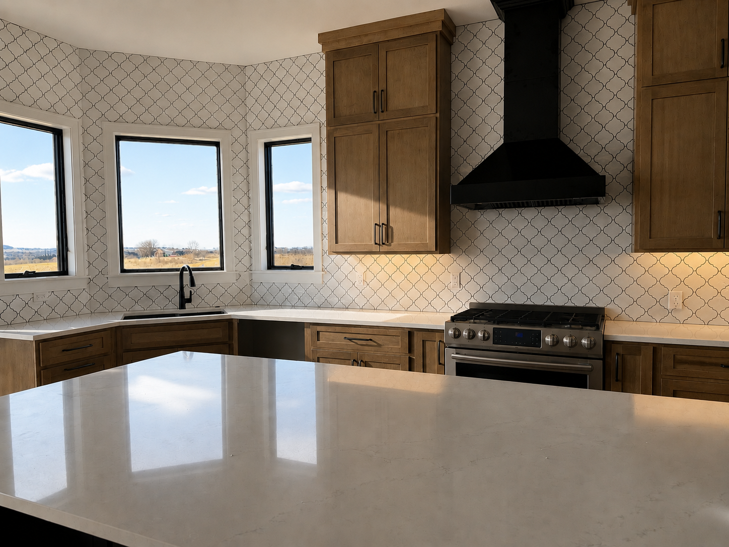 Modern kitchen with white patterned backsplash, wooden cabinets, black range hood, stainless steel stove, and a large white island in the foreground with a view of landscape through three windows.