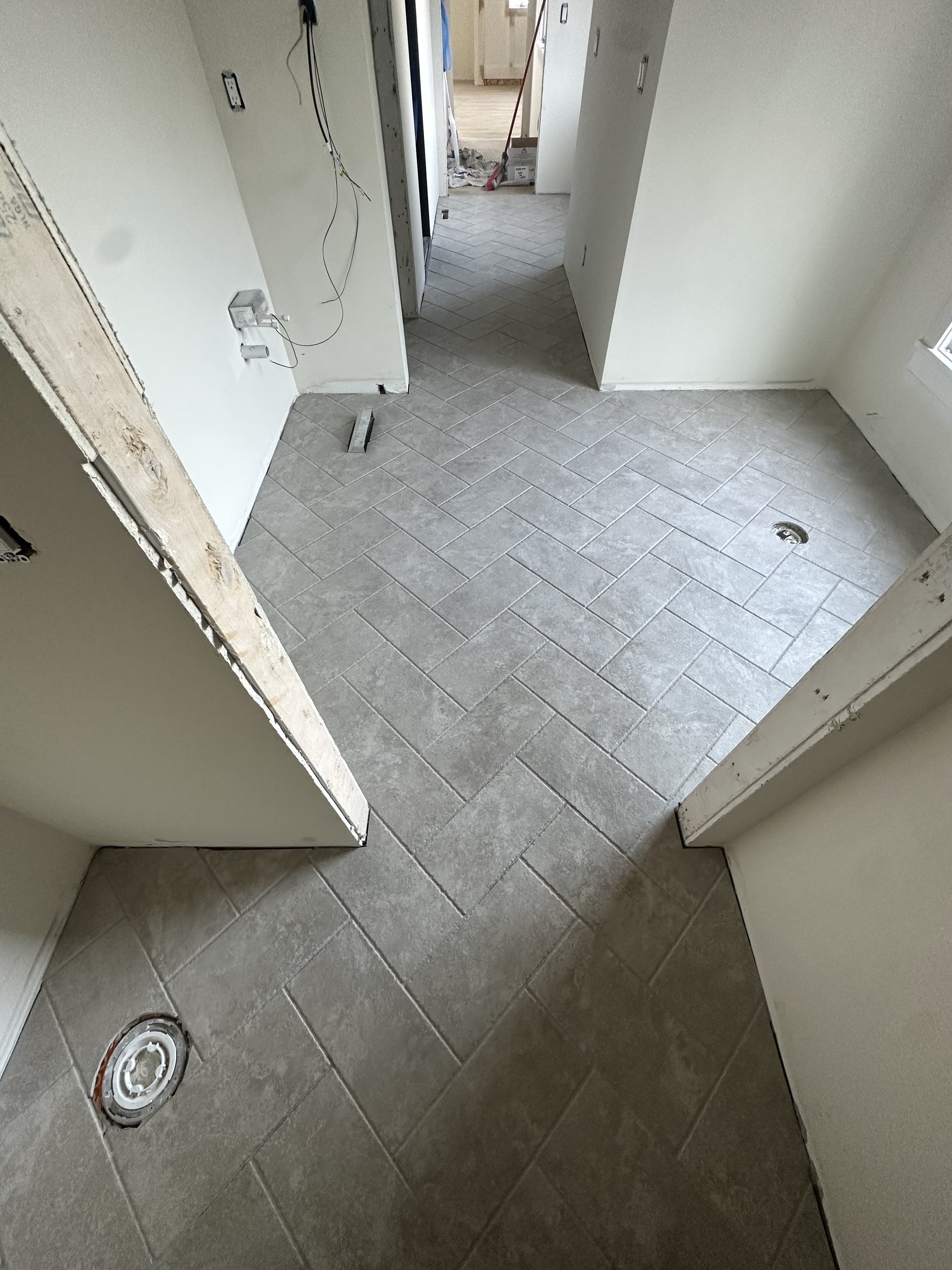 Interior view of a room under construction with beige tiled flooring, unfinished walls, and visible electrical wiring. A floor drain is visible, and there is an open doorway leading to another room with construction materials and tools.