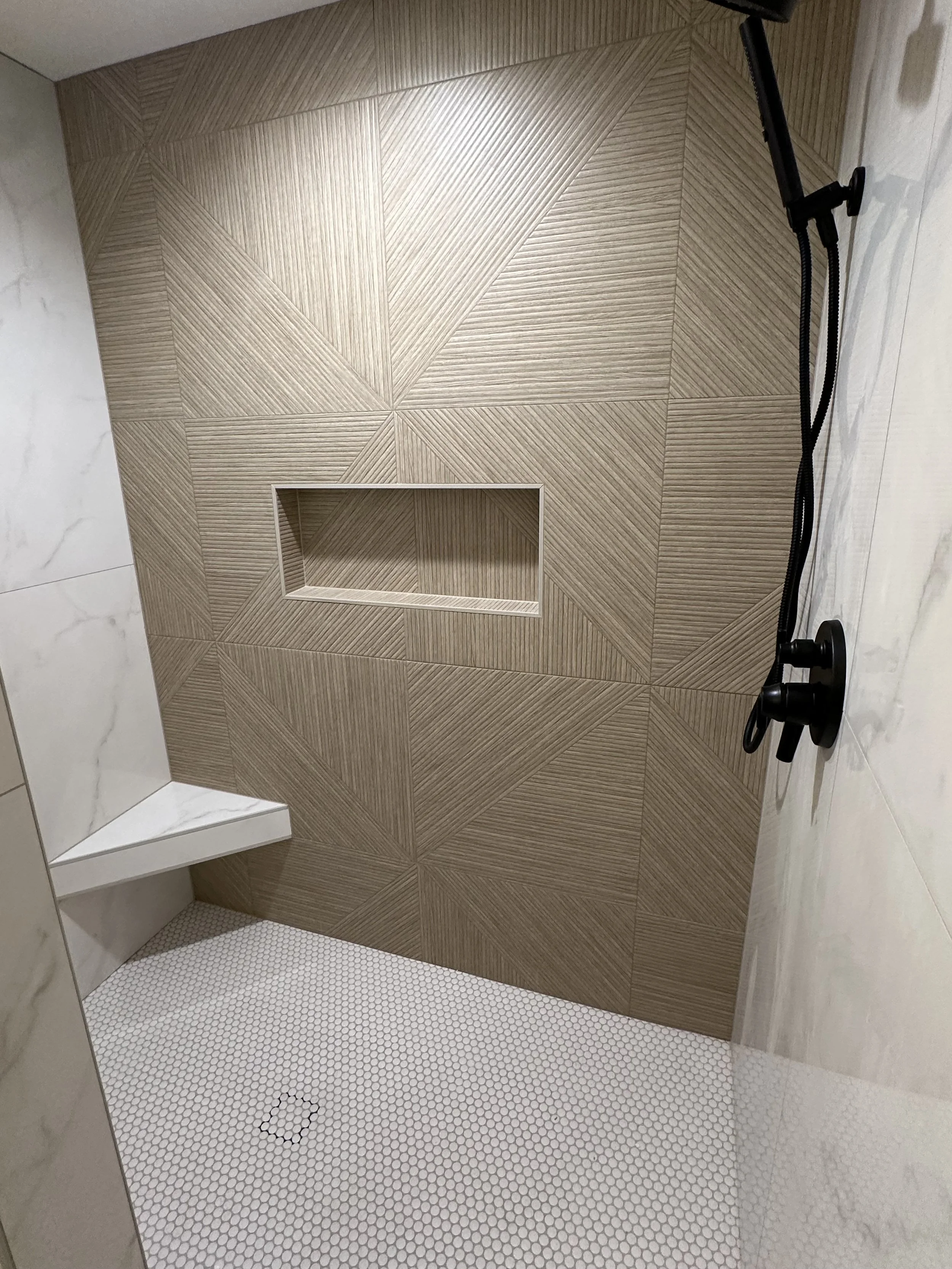 Bathroom shower corner with geometric wood tile walls, white hexagon tile floor, built-in niche, and black shower fixtures.