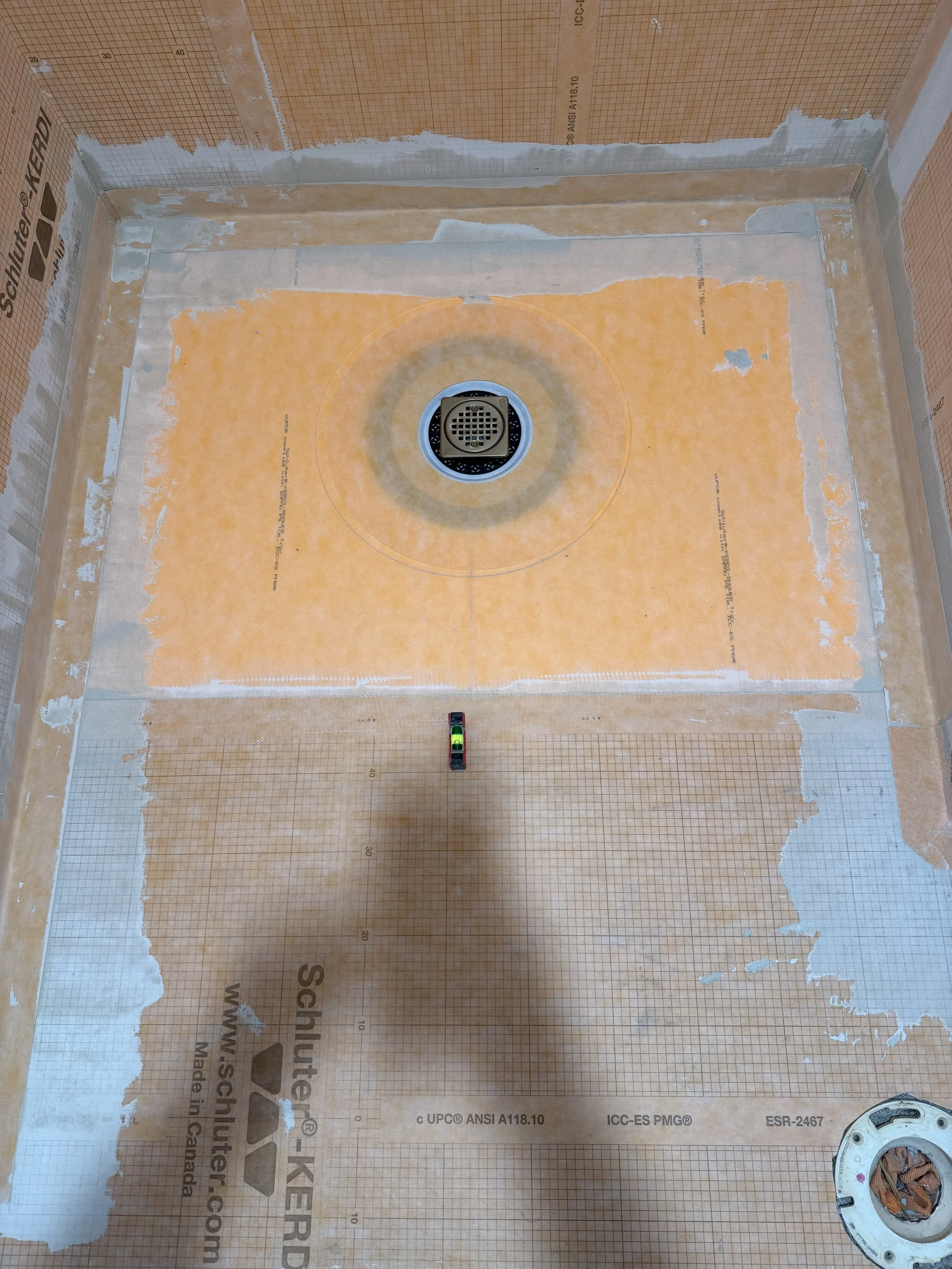 Bathroom floor with a drain pipe and moisture barrier membrane, showing torn edges and a level tool.