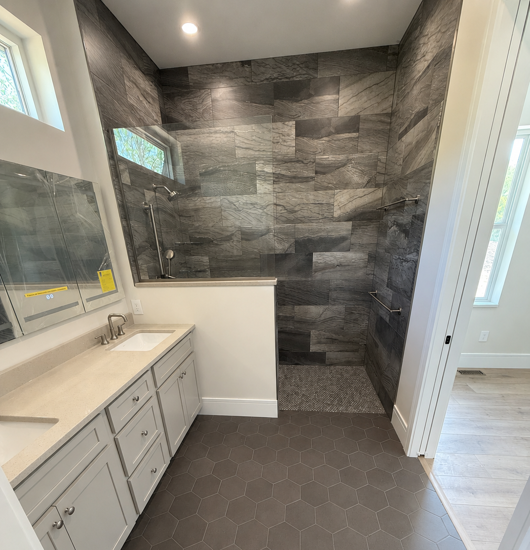 Modern bathroom with a shower area featuring dark gray marble tiles and glass door. Small vanity with a cream-colored countertop and white cabinets with silver knobs. Hexagonal brown floor tiles. Two windows providing natural light.
