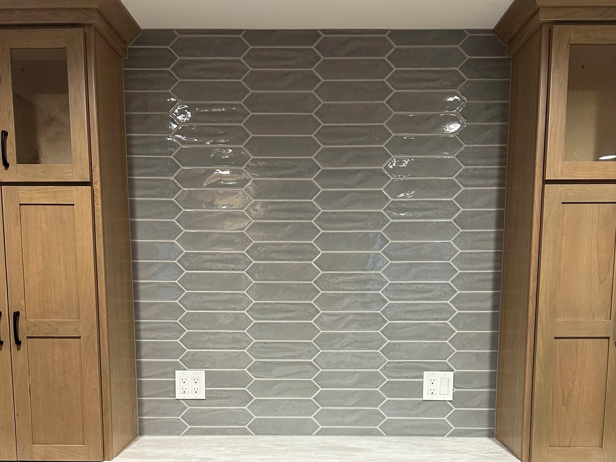 A kitchen wall featuring dark gray hexagonal tiles with white grout, flanked by two wooden cabinets on each side. There are two electrical outlets at the bottom of the wall.