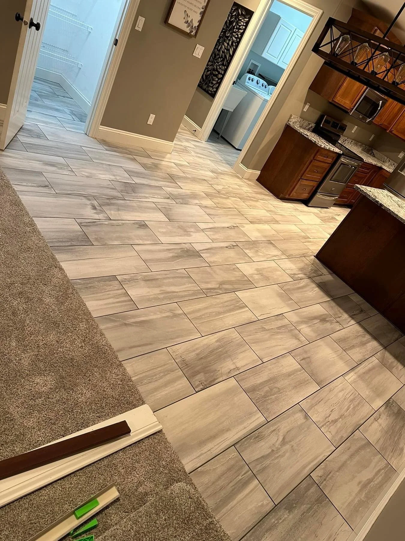 View of a kitchen with brown cabinets, granite countertops, and stainless steel appliances. Tiled floor with a creamy beige pattern. Part of a carpeted area is visible at the bottom left corner.