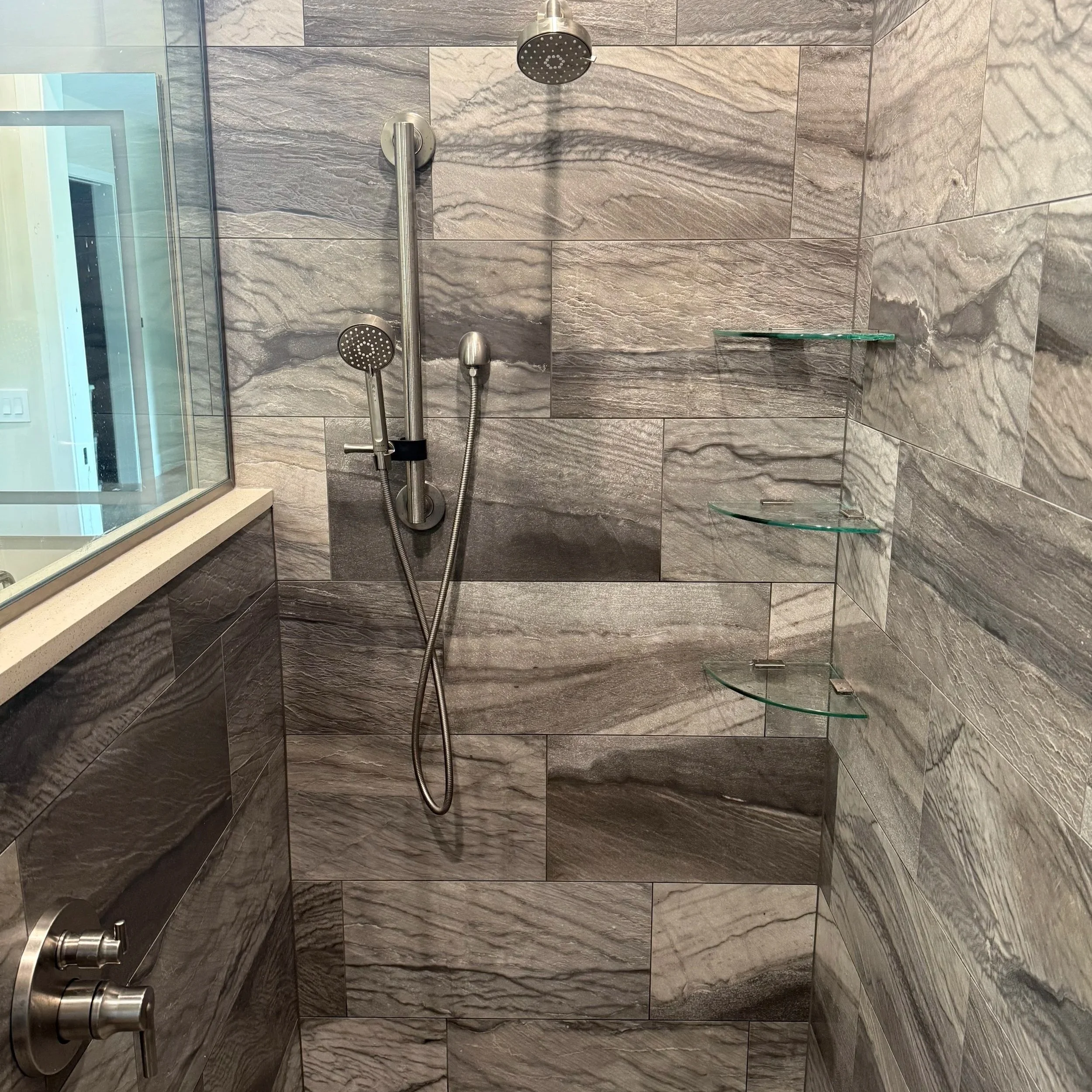 A modern shower with dark grey marble tiles, a fixed shower head, a handheld shower wand, a chrome faucet, and two glass corner shelves.