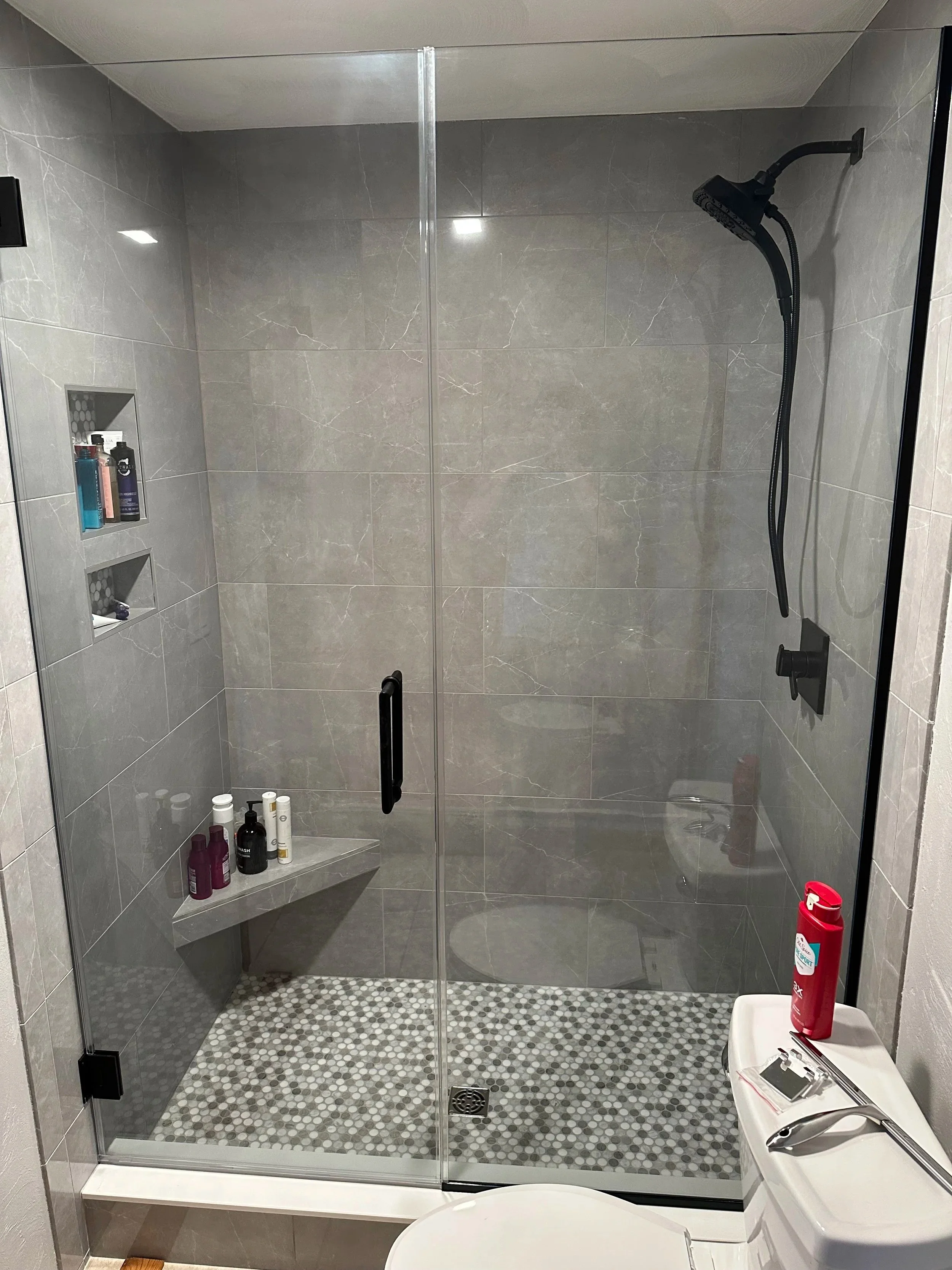 A modern walk-in shower with a glass door, grey marble tiles, built-in shelves holding shampoo and soap, a black showerhead, a small soap dish, a red fire extinguisher outside the shower, and a white sink with a faucet and a small mirror on top.