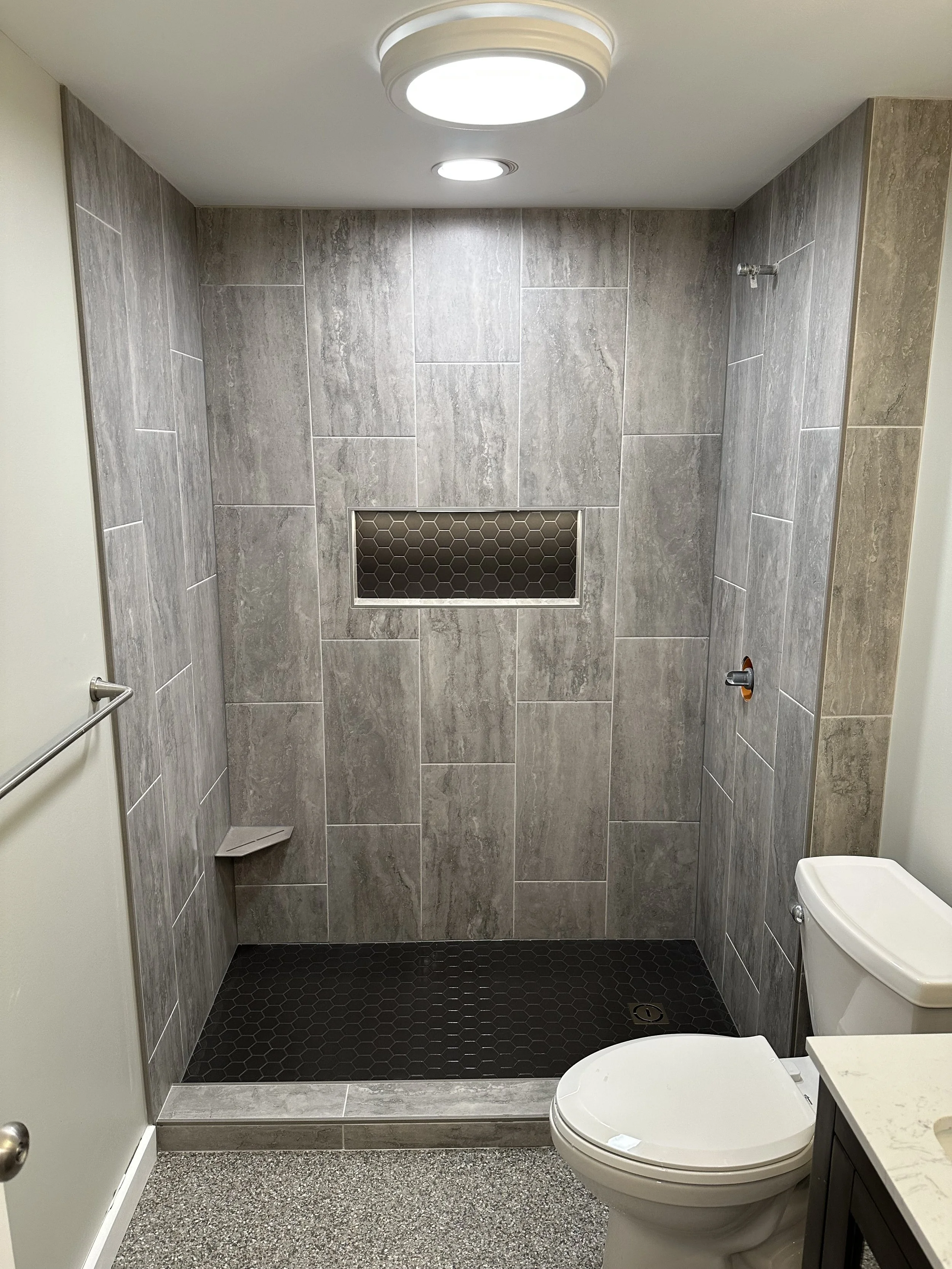 Bathroom with gray tiled walls, a toilet with a closed lid, a small countertop with a beige surface, a wall-mounted grab bar, a small corner shower area with hexagonal black tiles, a wall vent, a ceiling light, and part of a white bathtub.