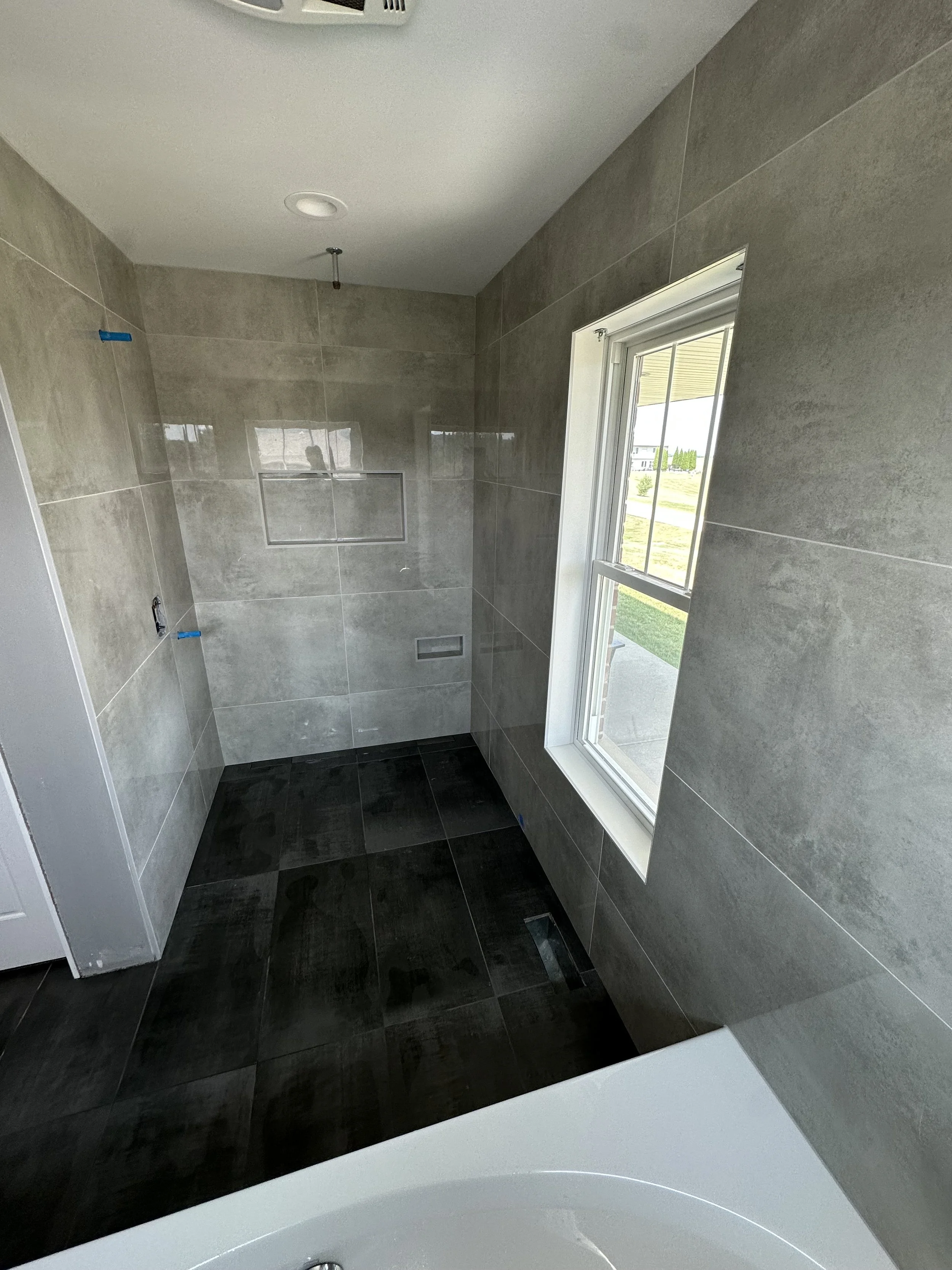 A bathroom under construction with gray tiled walls and black tiled floor, a window on the right, a ceiling vent, and an opening in the wall likely for a shower or bathtub.