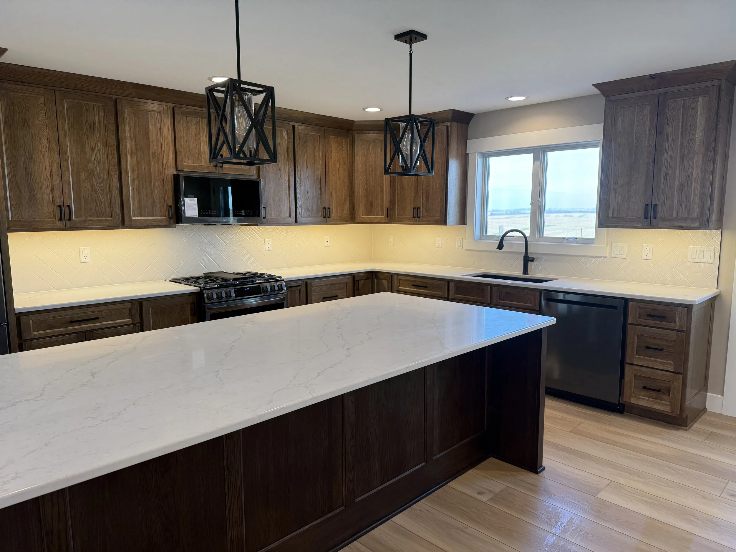 Modern kitchen with wooden cabinets, white marble countertop, black pendant lights, and a large window with a view of outdoor landscape.