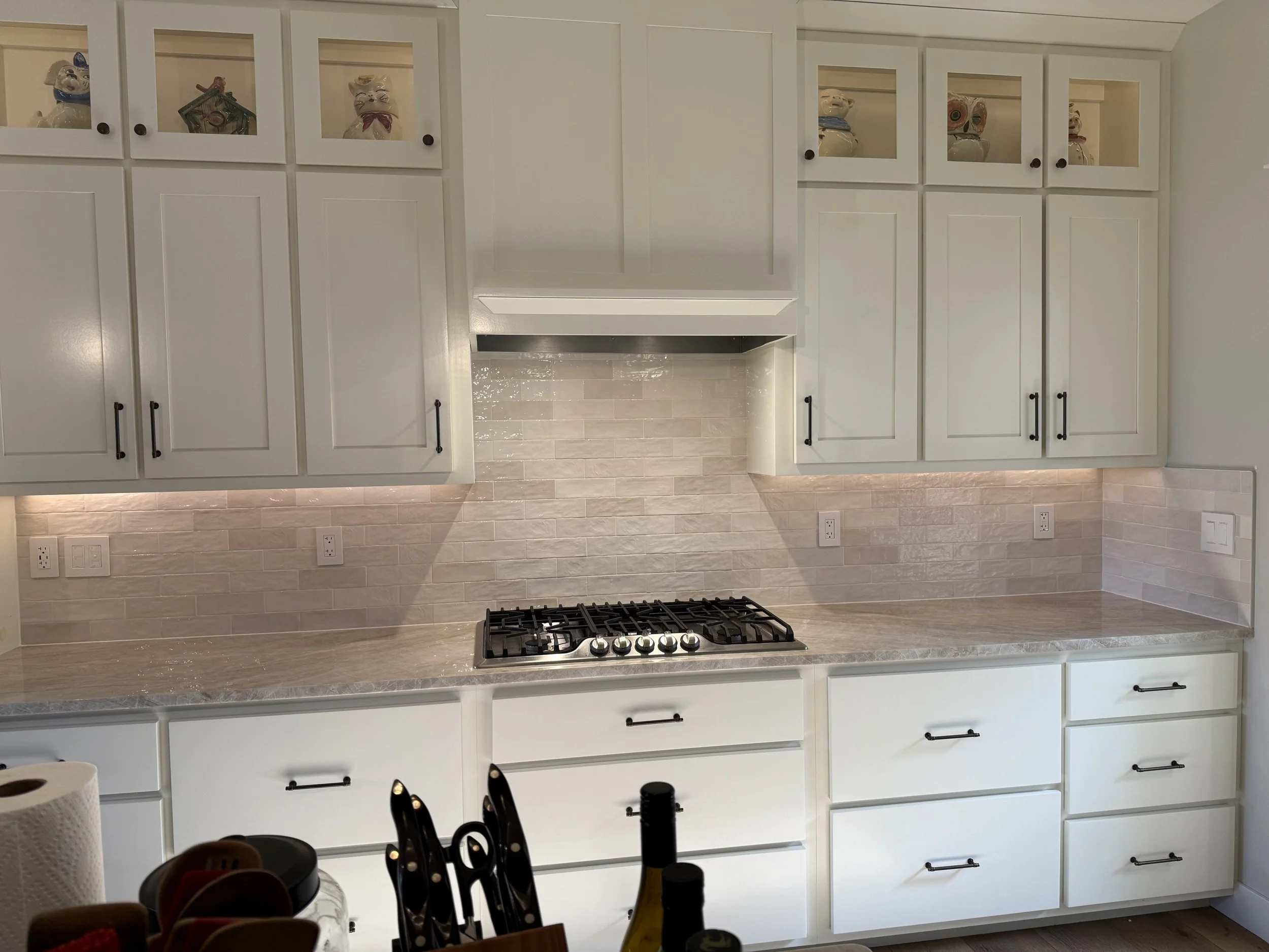 White kitchen cabinets with black knobs and glass-front upper doors decorated with pig-themed figurines, a stove with black grates, and a beige tiled backsplash.