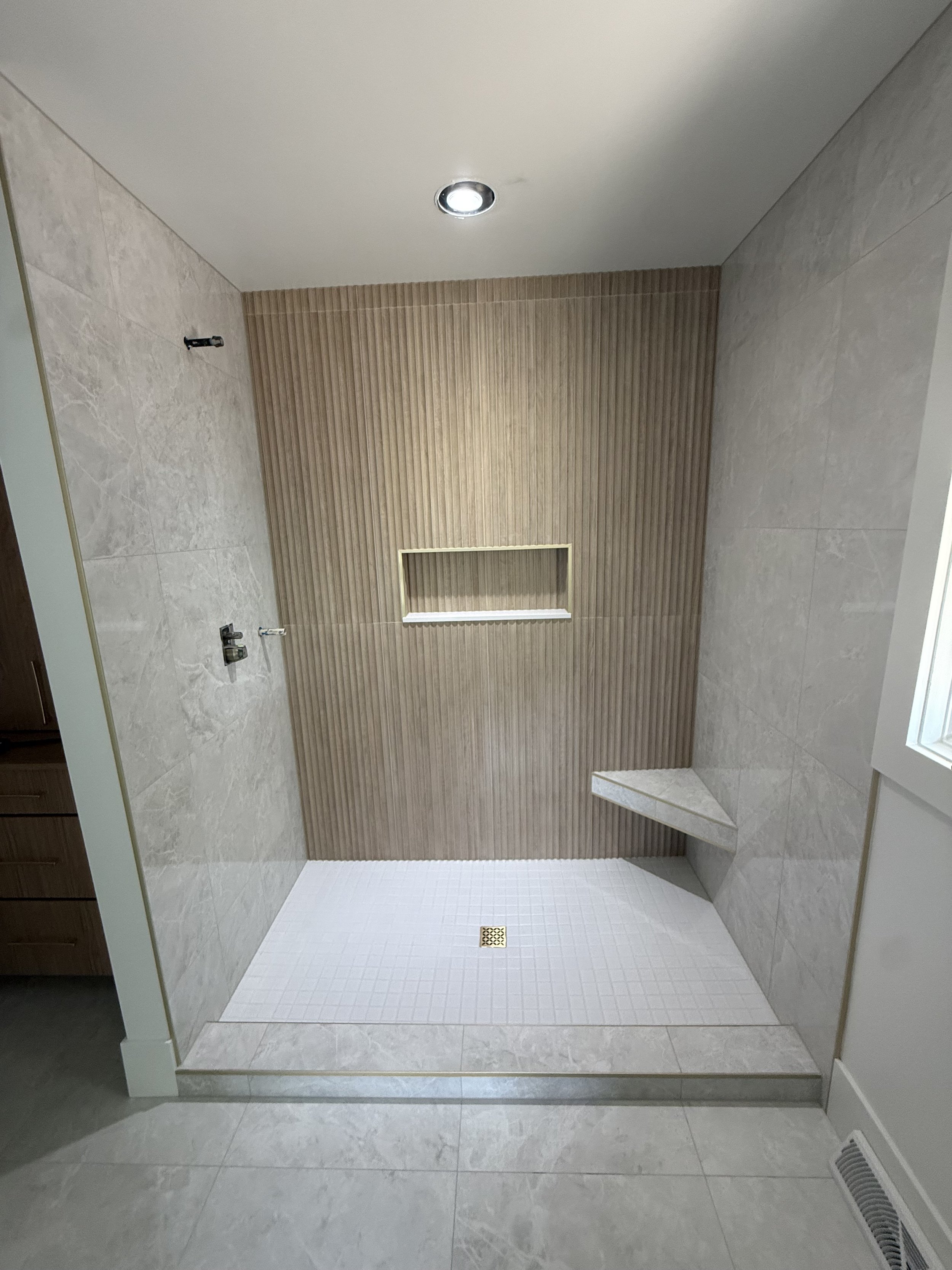 A bathroom shower area with beige tile and wood-paneled wall, a built-in niche, and a white tiled floor with a drain, no fixtures installed.